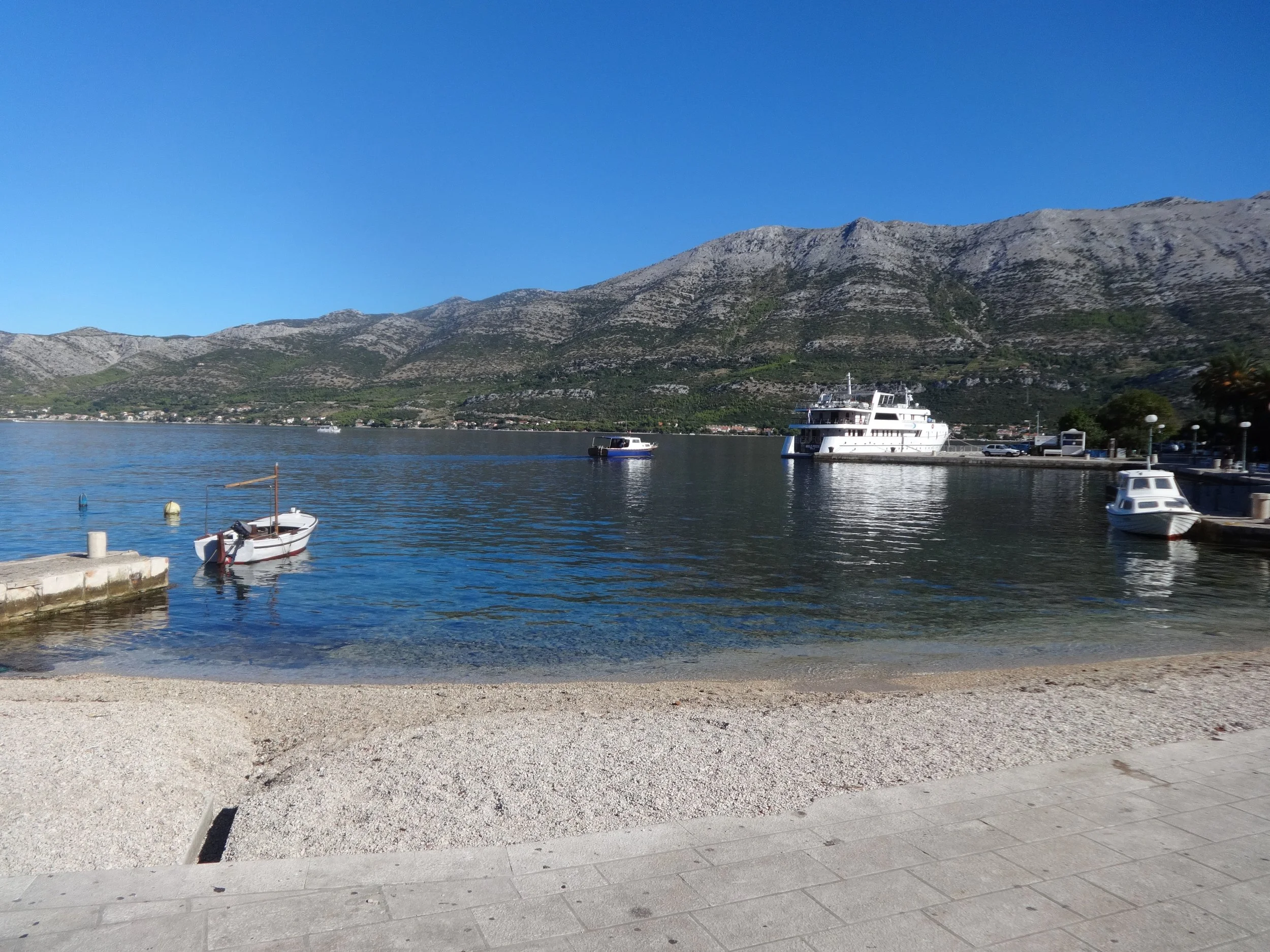 View of a mountain range with a water body in the foreground, boats docked along the shoreline, and a clear blue sky.