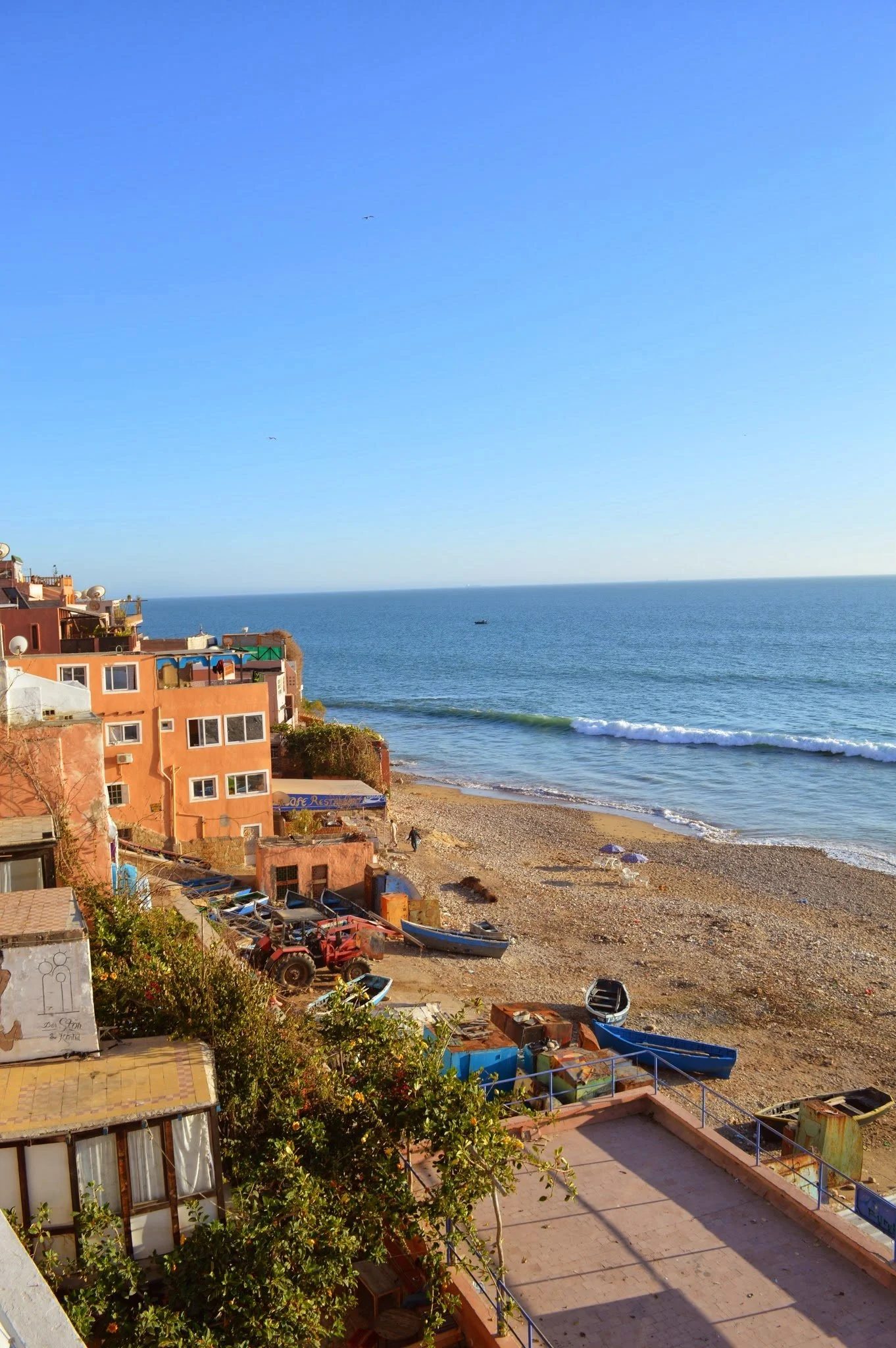 View of a beach with small waves, a boat in the distance, and a partly sunny sky. In the foreground, there are colourful buildings, boats, and a patio area with shrubs and outdoor furniture.