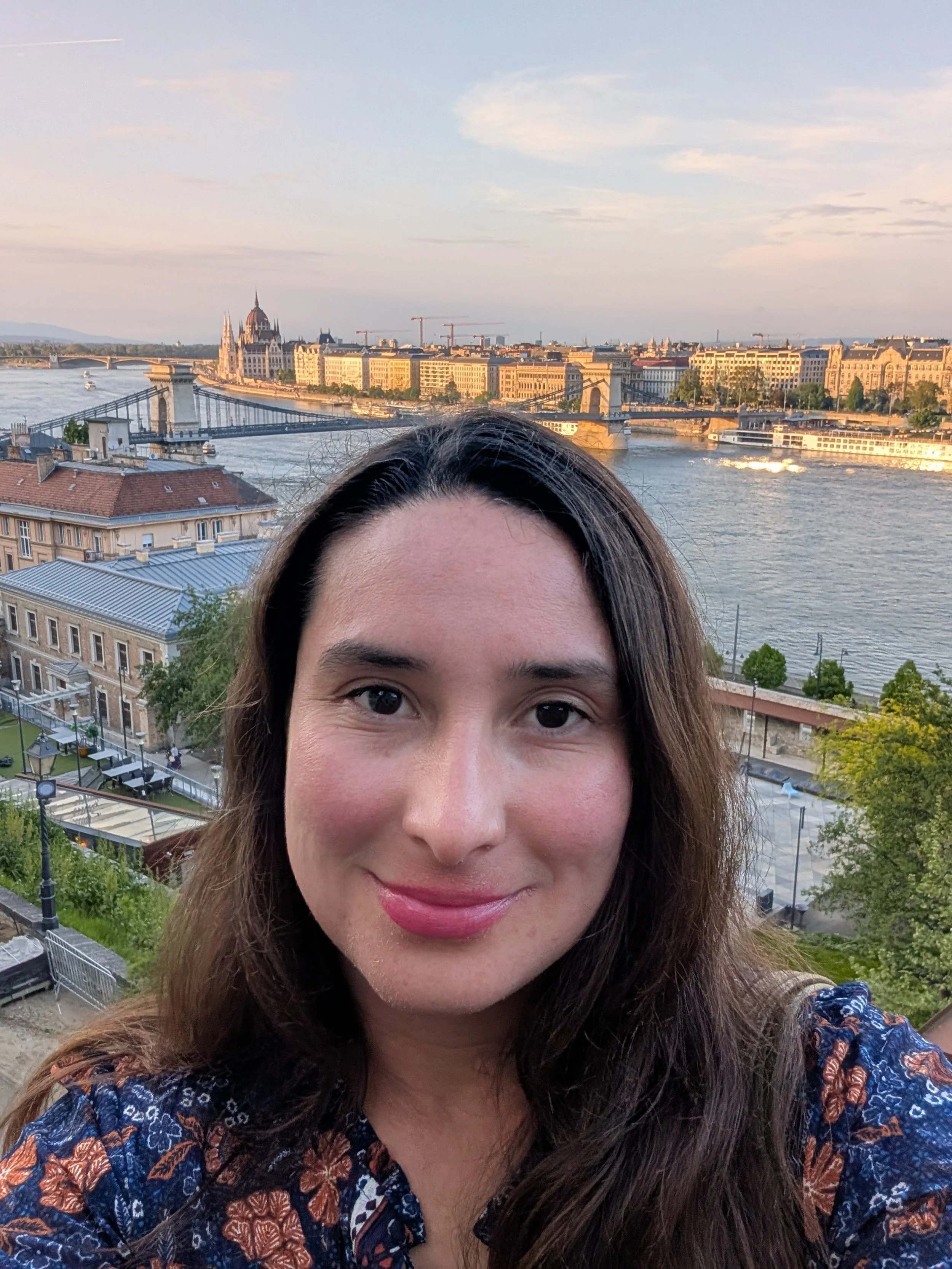 A woman with long brown hair smiling outdoors with a river and city skyline in the background, including a bridge and historic buildings.