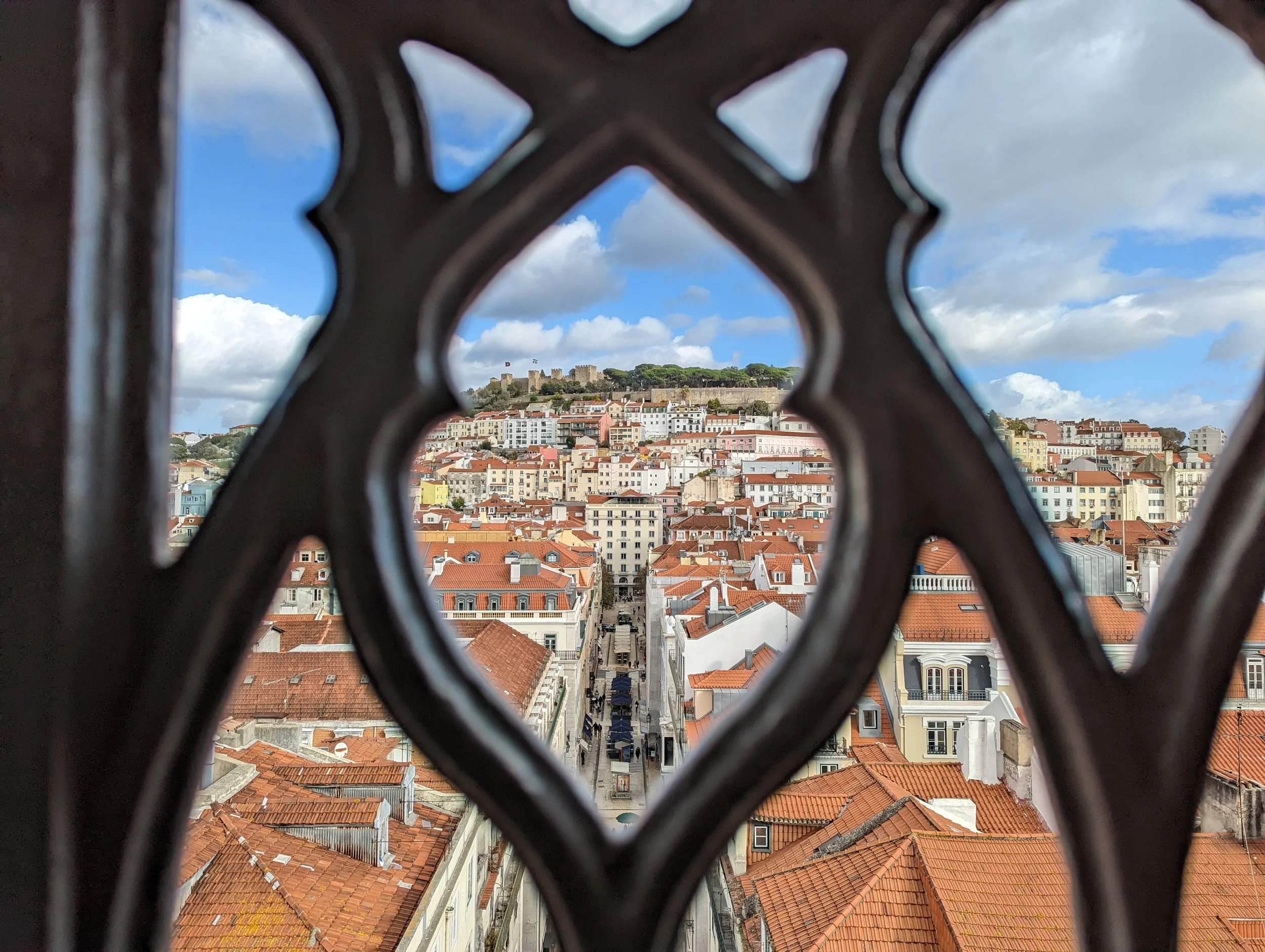 View of a cityscape with red-tiled roofs taken through decorative ironwork. There are buildings, a street, and a hilltop with a historic castle and flags in the background under a partly cloudy sky.