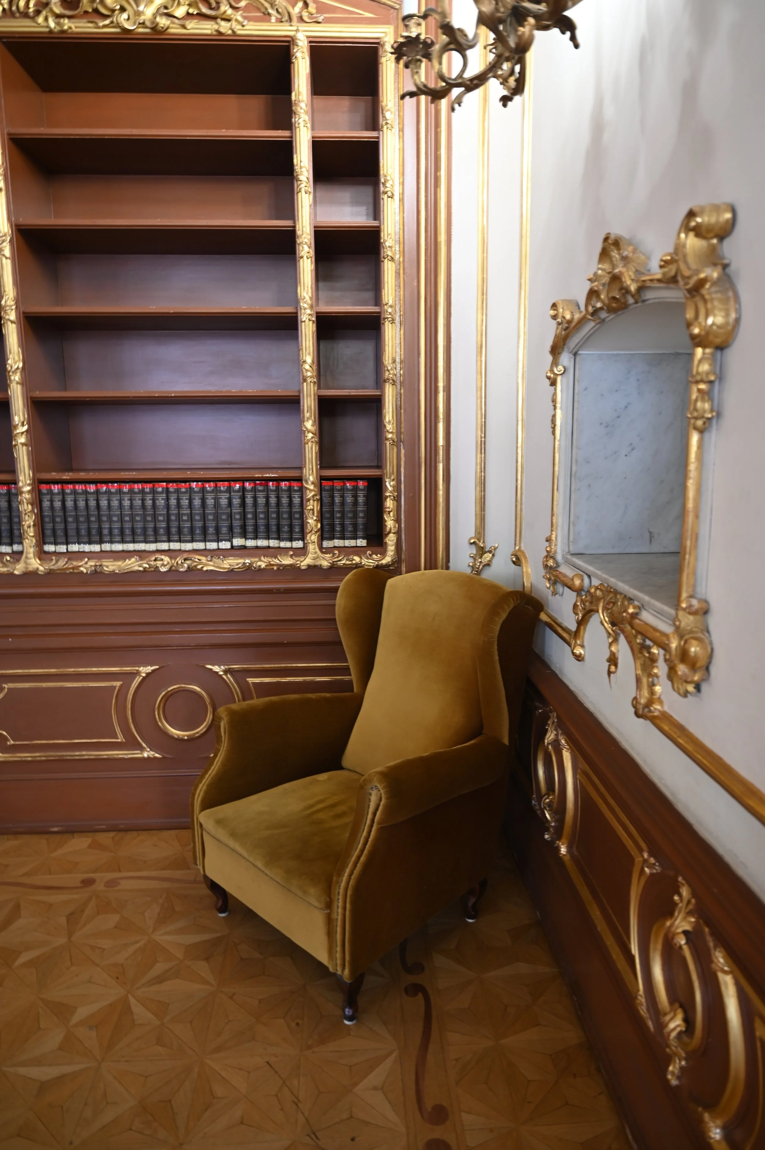 A gold ornate bookshelf filled with books, a matching gold framed mirror on the wall, a vintage chandelier, and a plush olive-green armchair, all in a luxurious room with parquet wooden flooring.