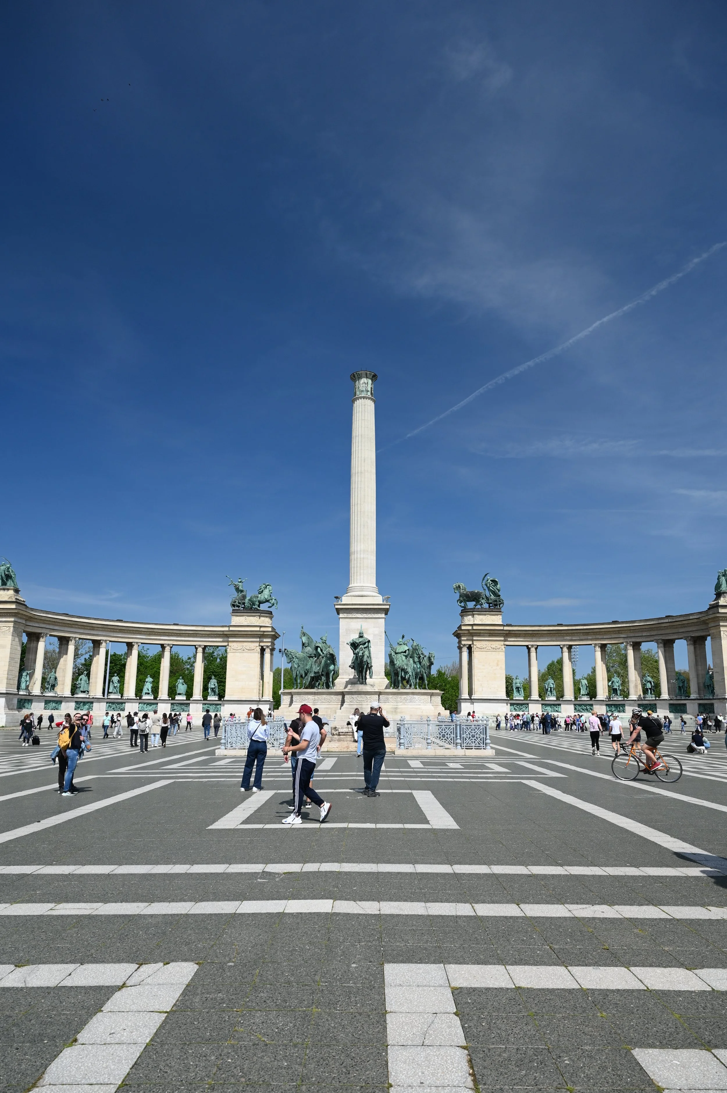 People walking and cycling in front of a large monument with a tall column and statues on a circular colonnade, under a blue sky.