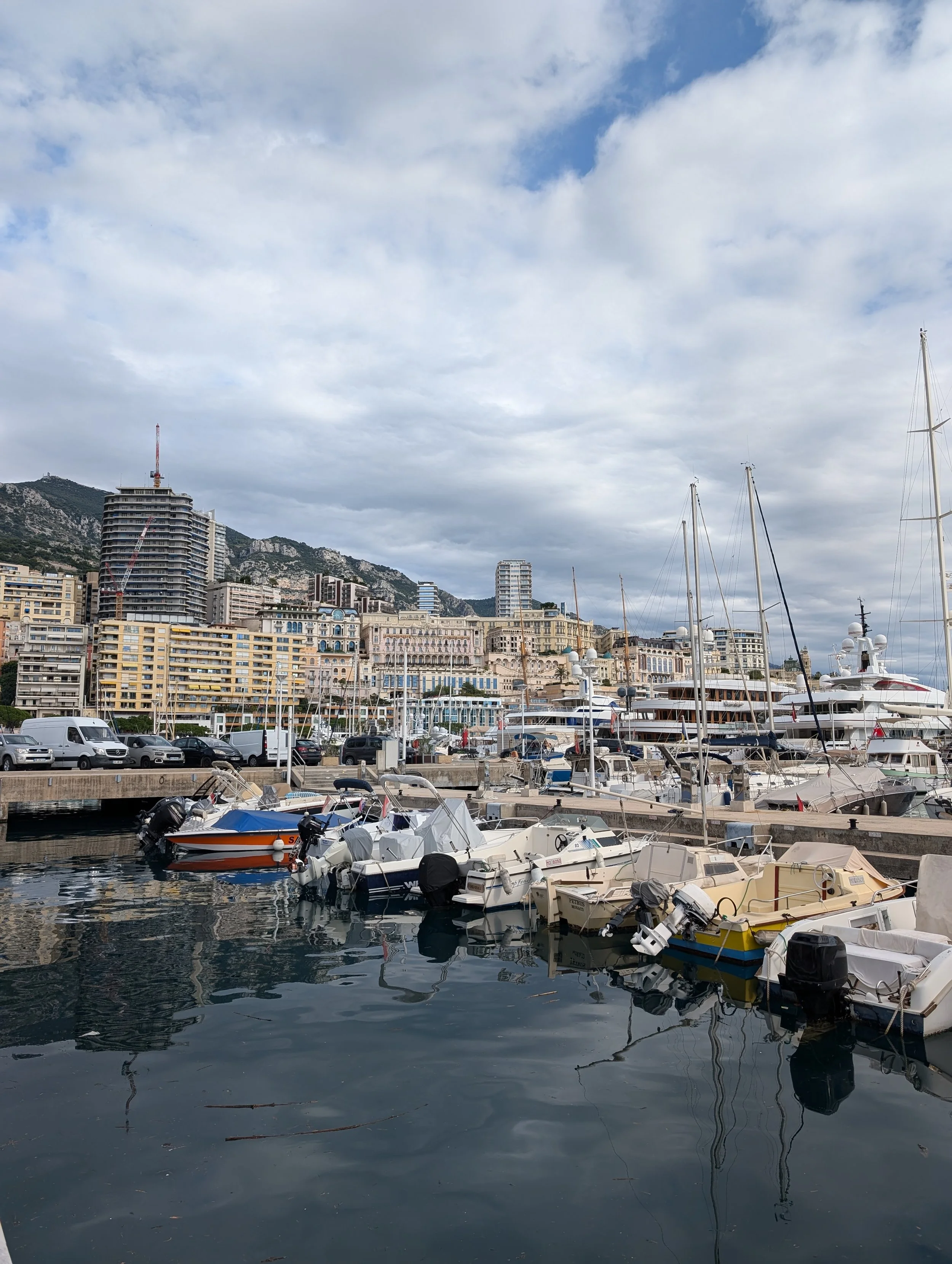 A marina with numerous boats docked in the water, with city buildings and mountains in the background under a partly cloudy sky.