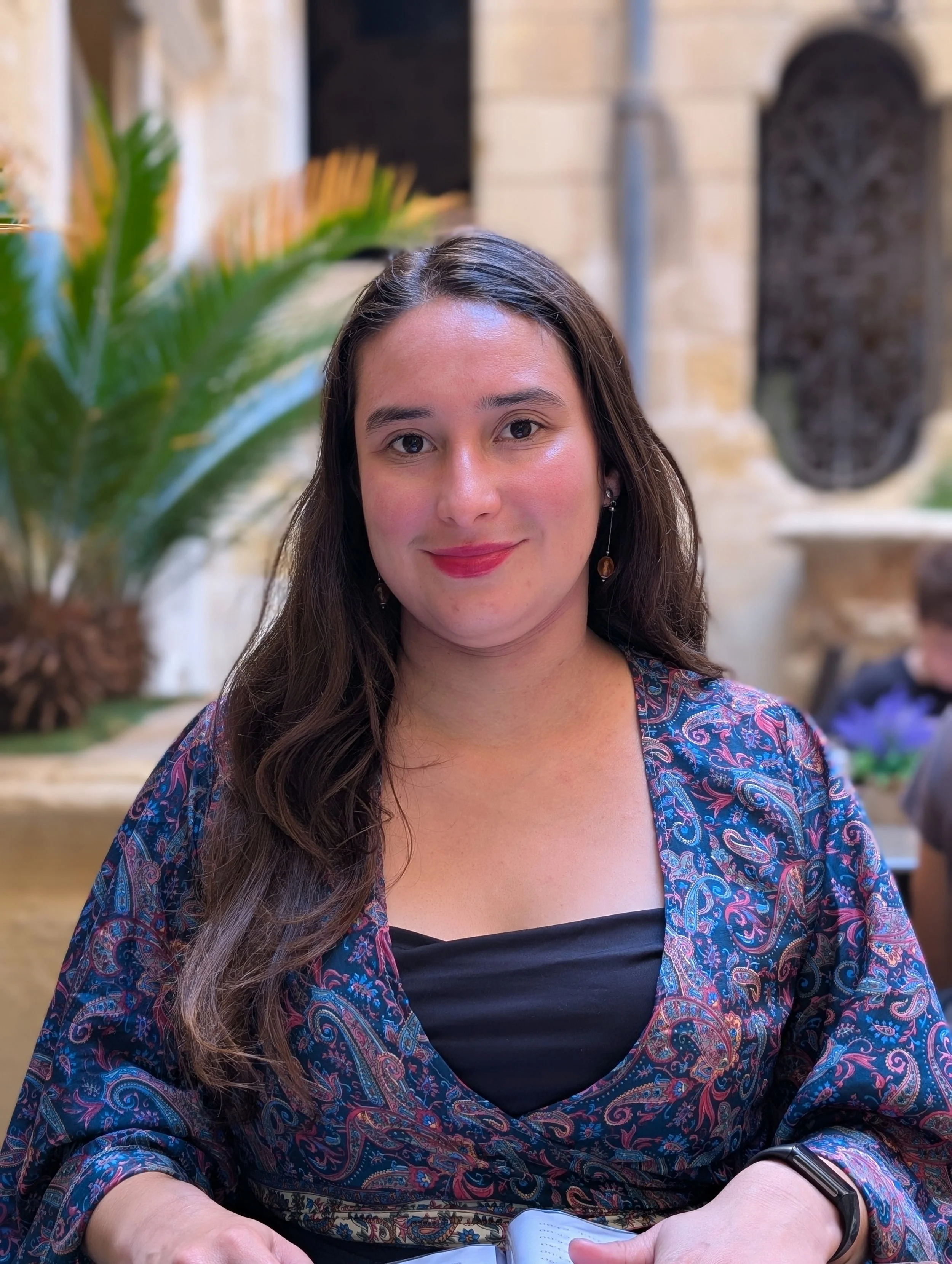 A woman with long brown hair wearing earrings, a black top, and a patterned jacket, sitting outdoors. She is holding an open notebook or journal, and a smartwatch on her left wrist. The background features a stone wall with decorative elements and gr