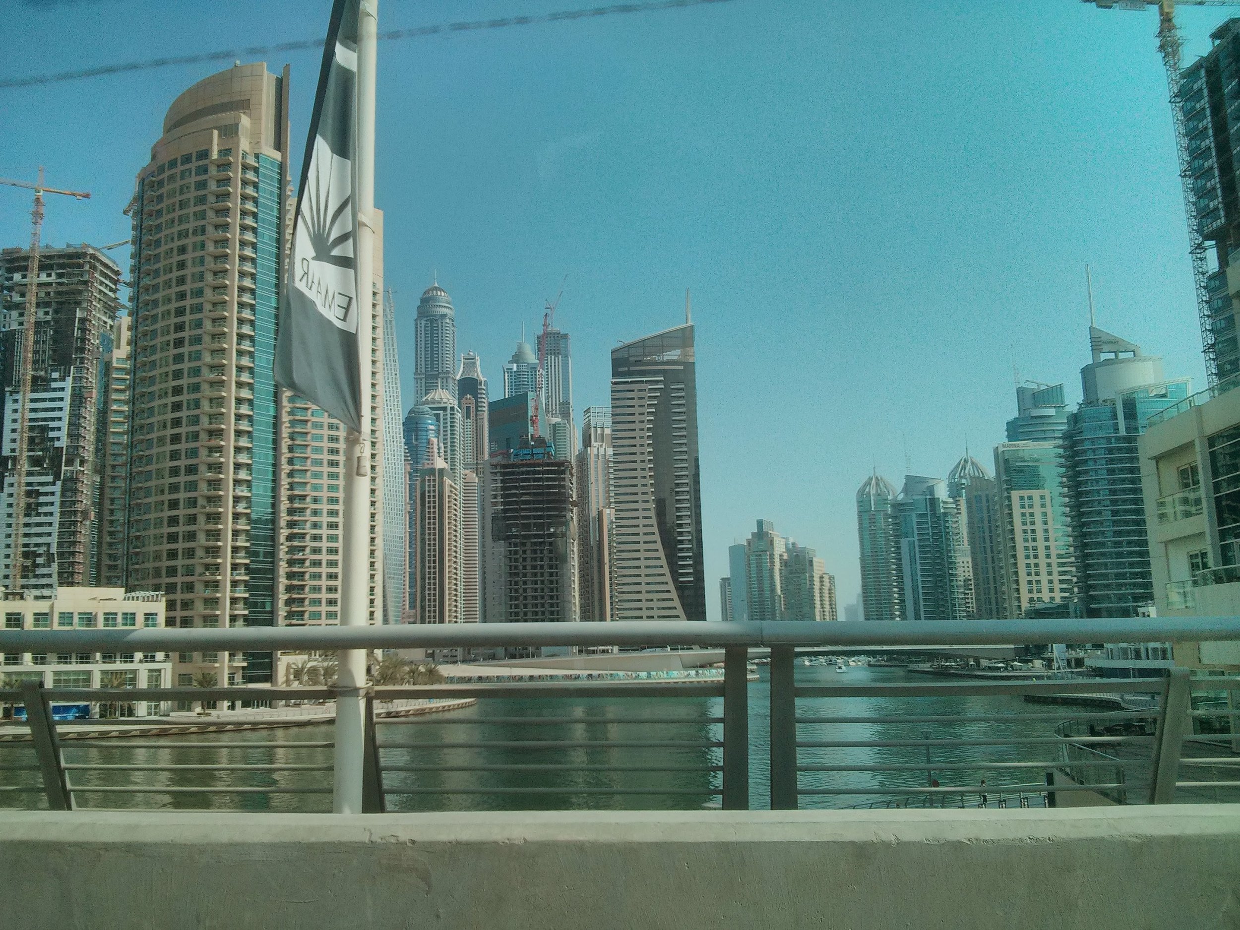 View of a city skyline with modern high-rise buildings, a waterway, and construction cranes under a clear blue sky.