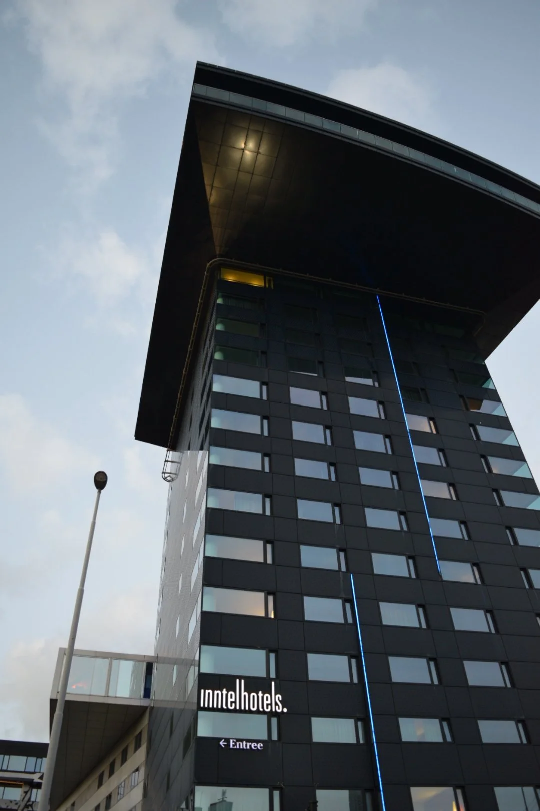 Tall modern building with a distinctive overhanging top, black exterior, and illuminated blue vertical line on facade, with 'innothotels' signage at the bottom and an entrance sign pointing left.