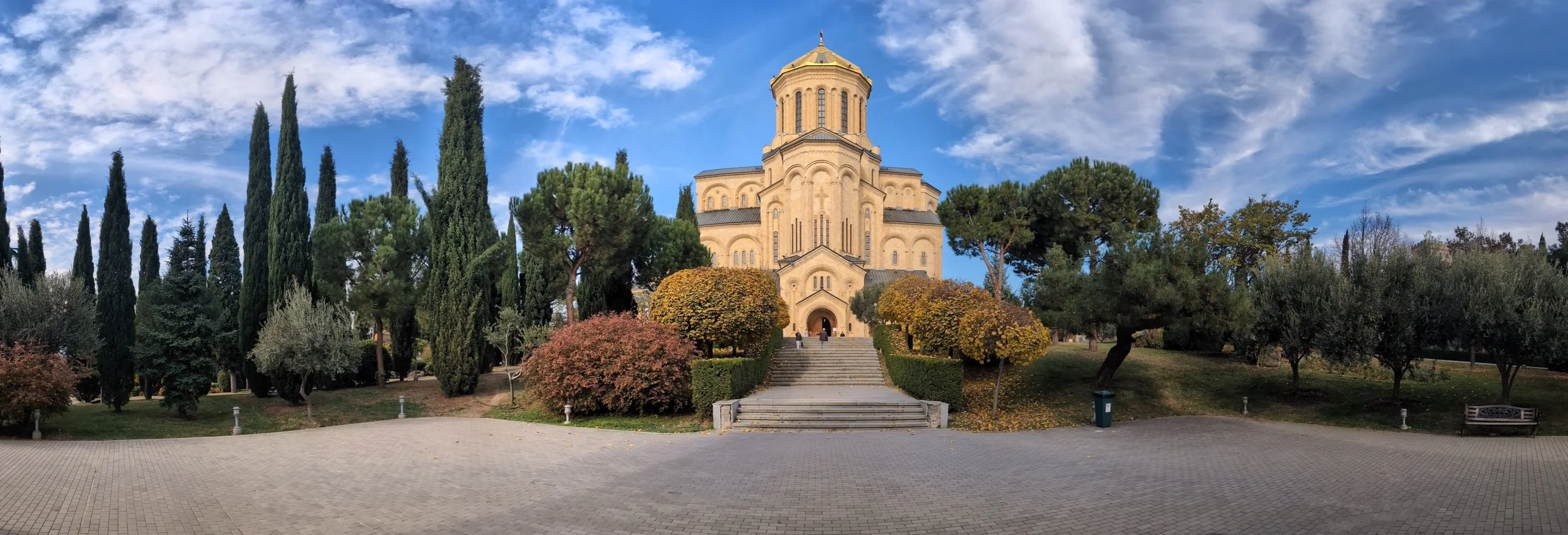 A historic church with a prominent central tower, surrounded by trees with fall foliage and a paved courtyard with stairs leading to the entrance.
