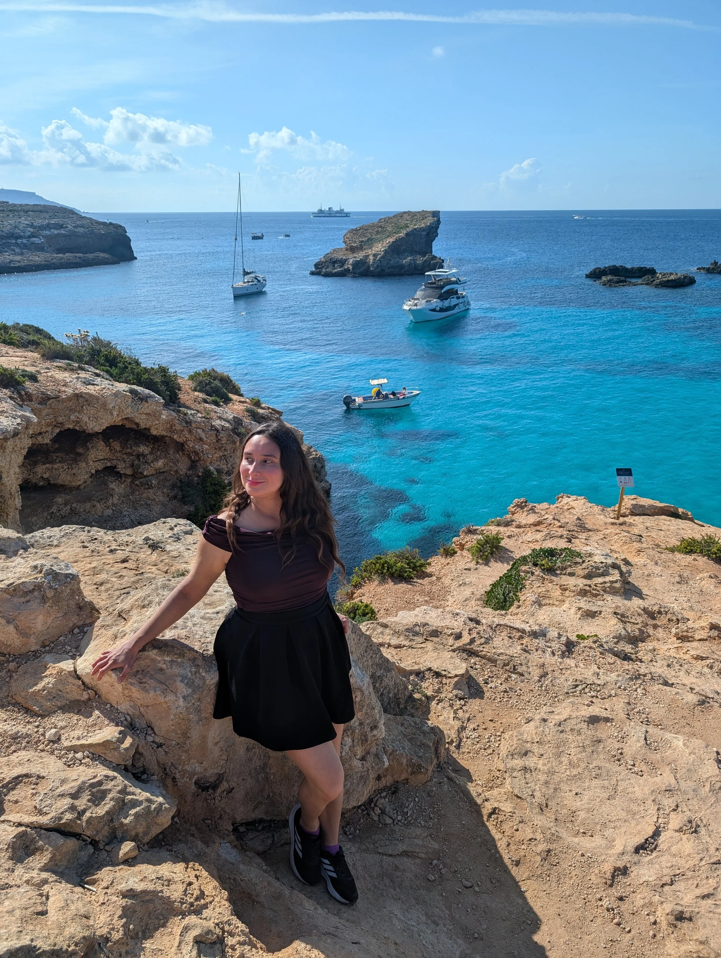 A woman standing on rocky terrain near the coast with blue sea water, boats, and a large rock formation in the background.