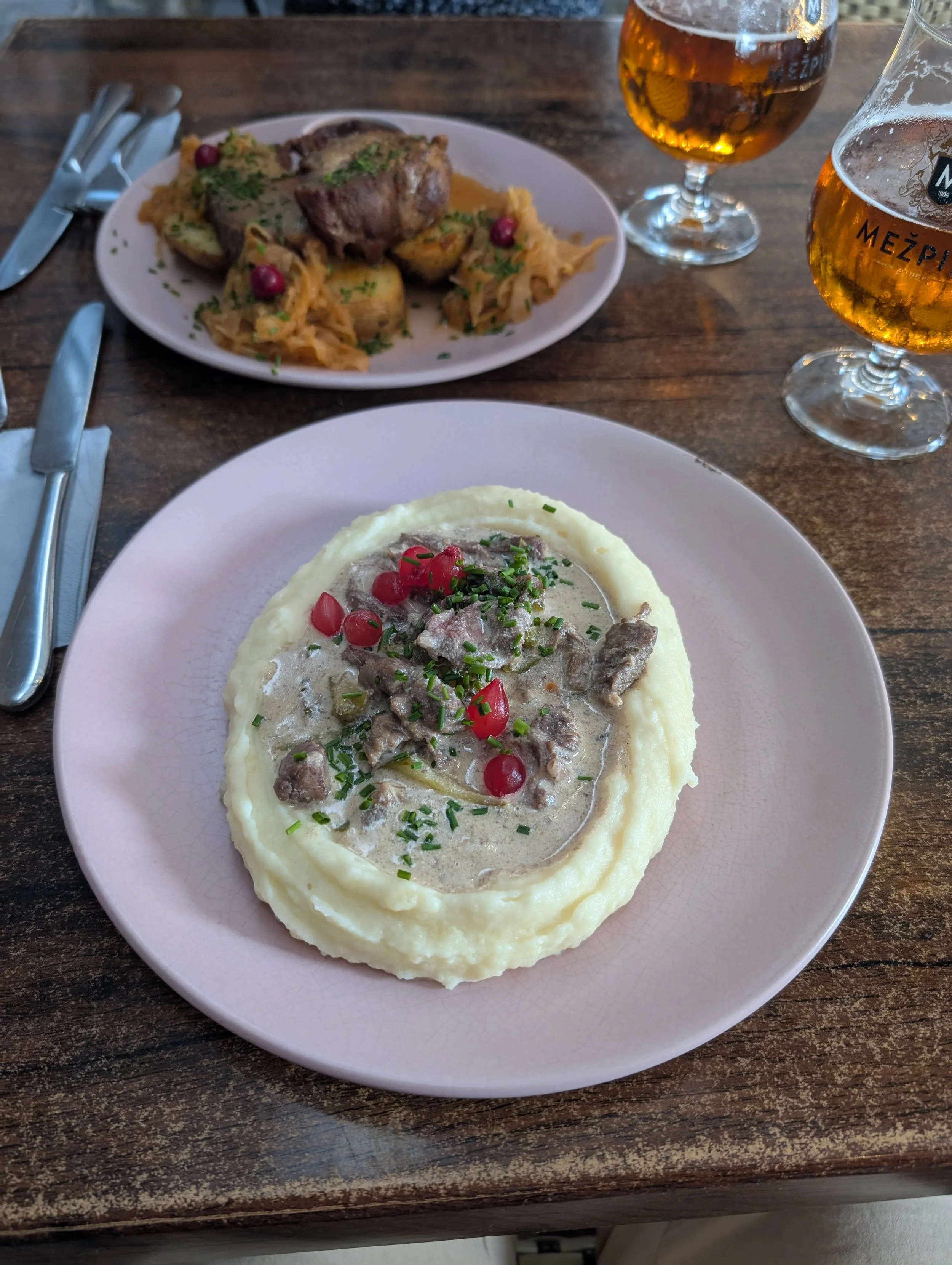 A plated meal of creamy meat stew garnished with red berries and green herbs, served with a side of mashed potatoes or buttered potatoes, on a pink plate. Two glasses of beer are in the background on a wooden table.