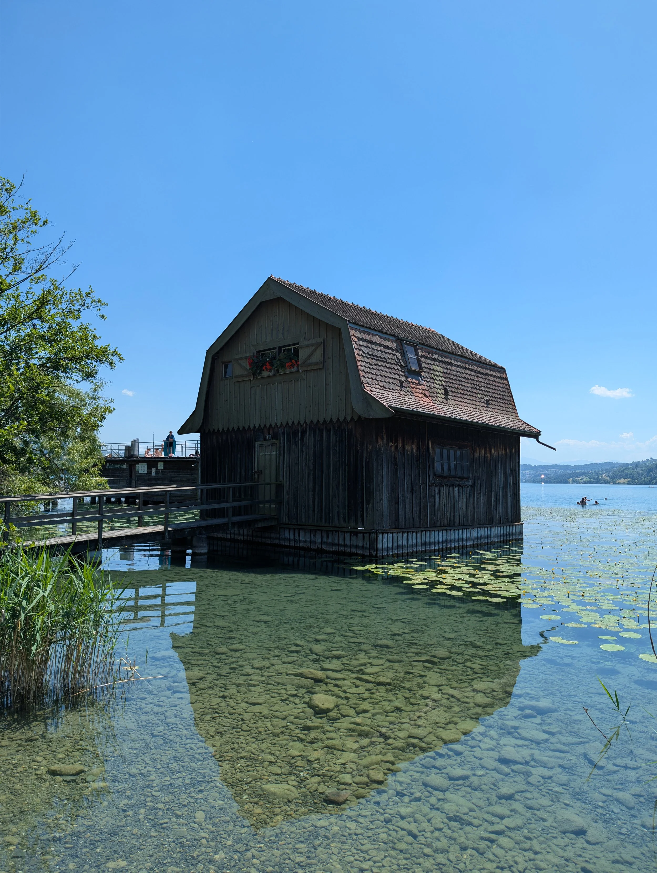 A rustic wooden house on the edge of a lake with a small deck and railing, under a clear blue sky, surrounded by greenery and distant mountains.