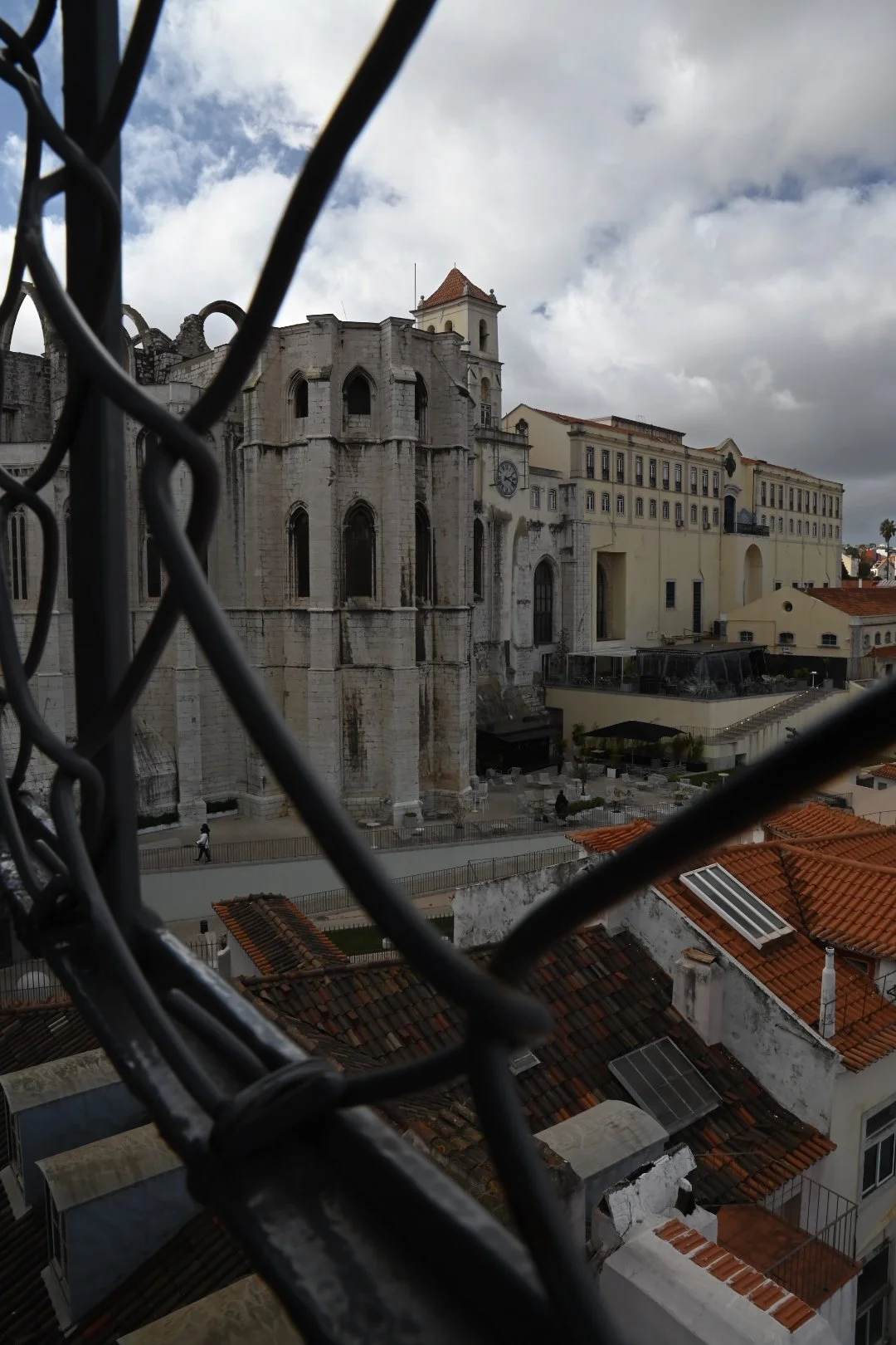 View of an old stone church or historic building with a clock and bell tower, seen through a black metal fence. Surrounding buildings have red-tiled roofs; the sky is cloudy.