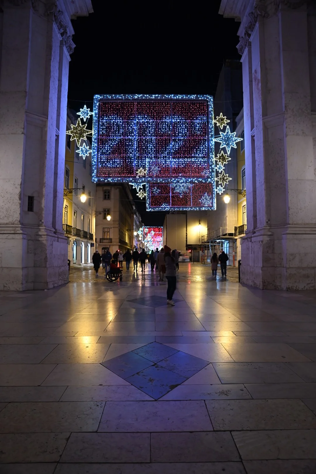 Night view of a street decorated with colorful Christmas lights and illuminated star-shaped decorations hanging overhead, with people walking around and looking at the displays.