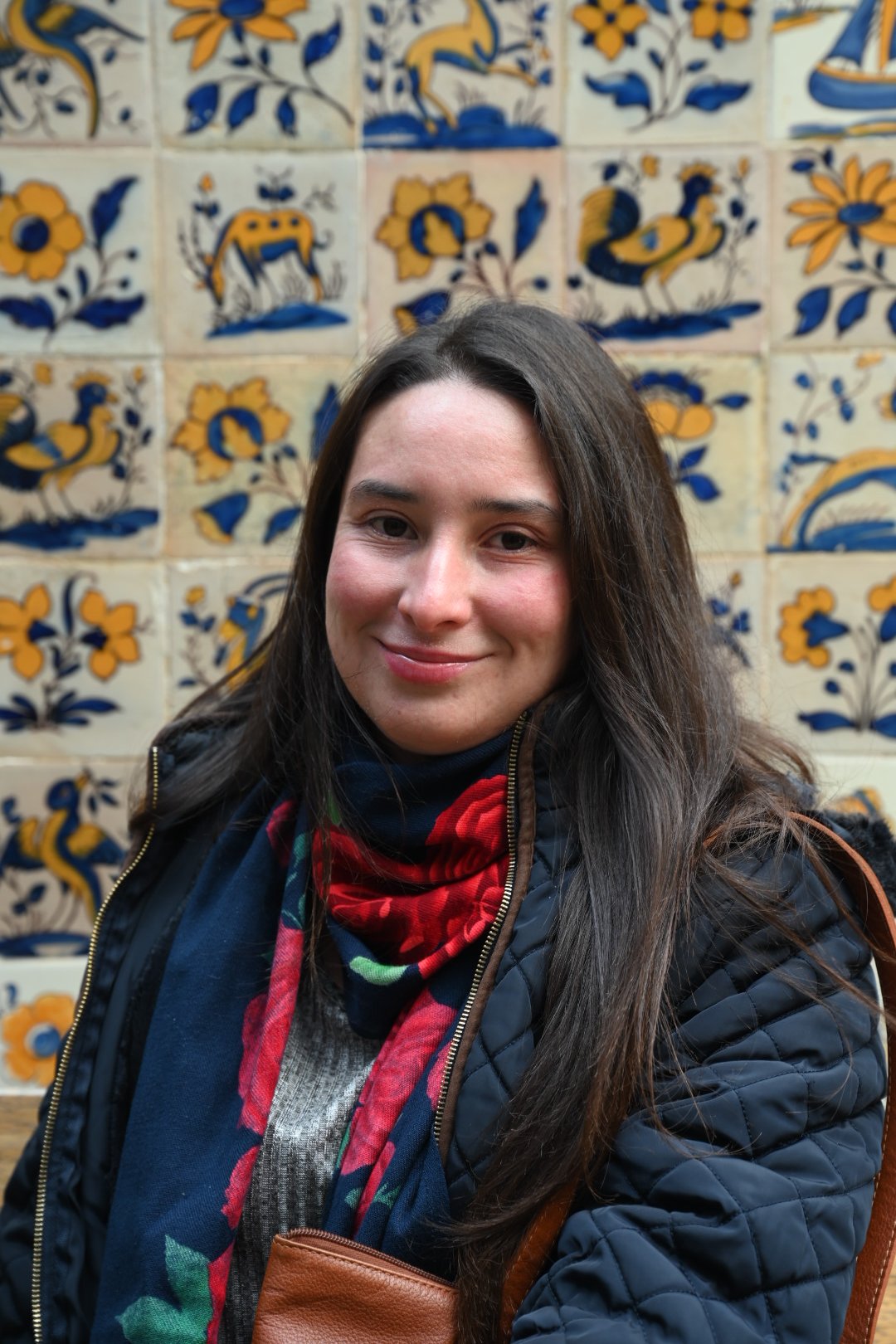 A young woman with long dark hair, smiling, standing in front of a decorative tiled wall with a floral pattern in blue, yellow, and white.