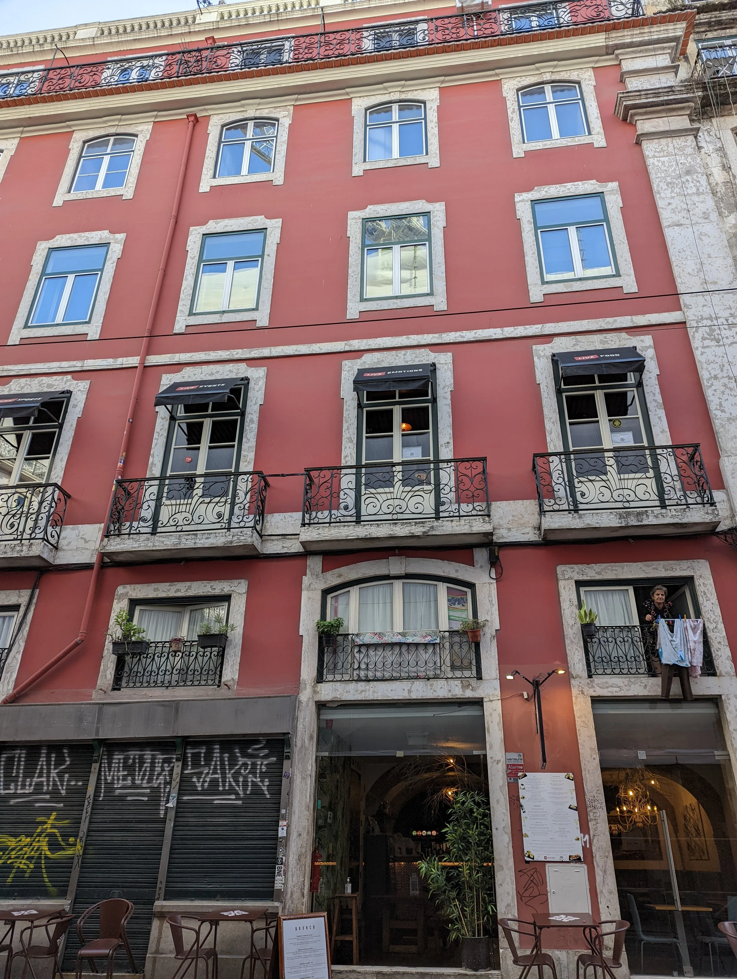 A three-story building with pink walls and white stone accents around the windows. The ground floor has a restaurant with open seating, tables and chairs outside, and a menu stand near the entrance. There are balconies with wrought iron railings on t