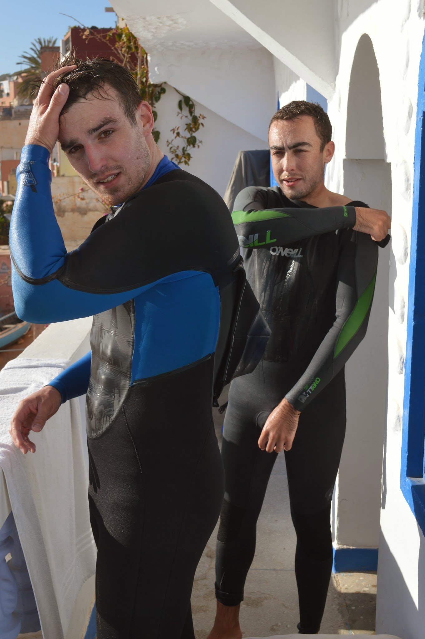 Two young men in wetsuits stand on a balcony or terrace, with a white wall and blue trim behind them. One man touches his head while the other looks into the distance. The scene appears sunny and warm.