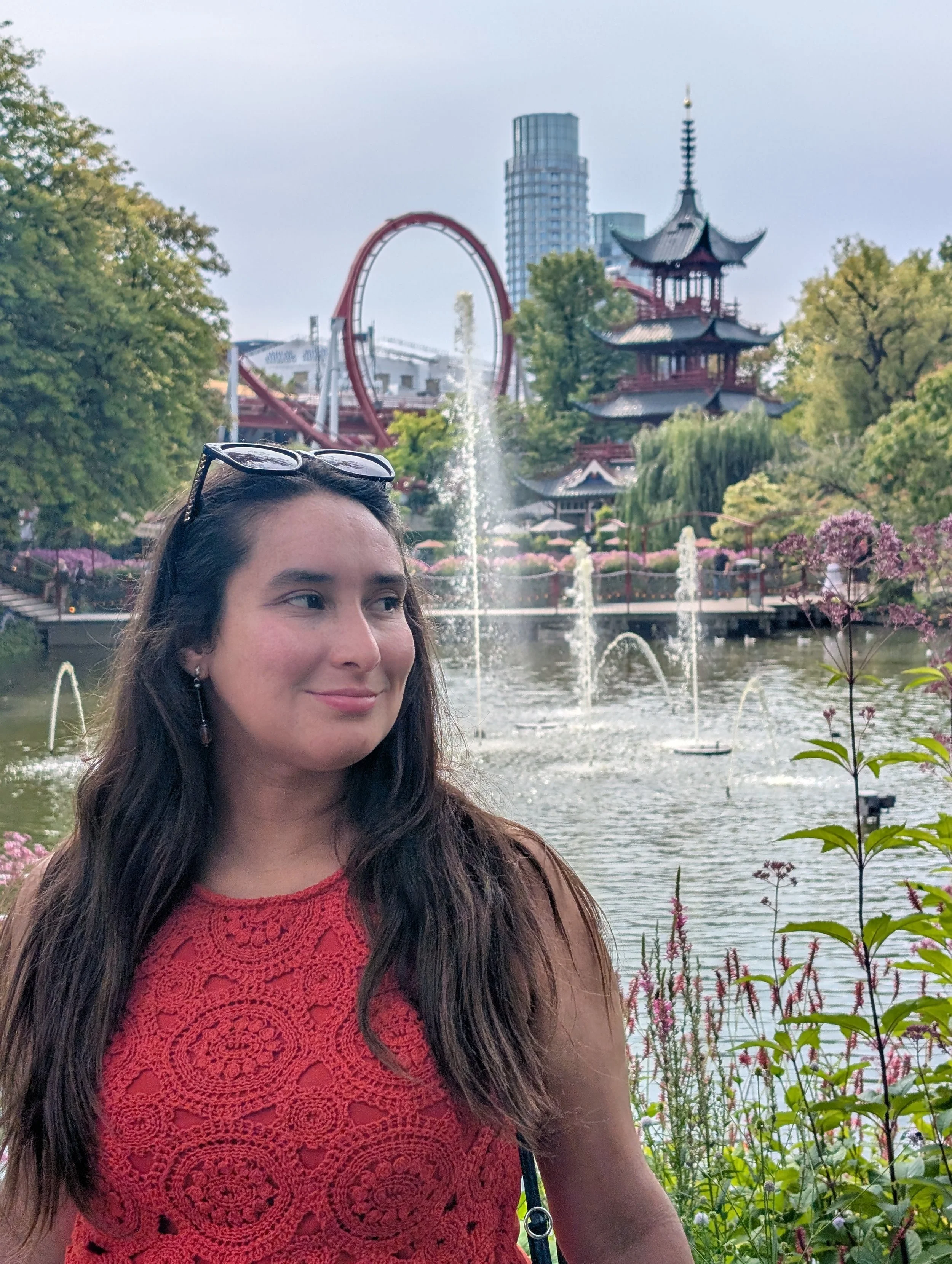 A woman with long dark hair and sunglasses on her head stands in front of a pond with fountains. Behind her are trees, flowering plants, a multi-tiered pagoda-style building, and a roller coaster with a red loop. Tall buildings are visible in the bac