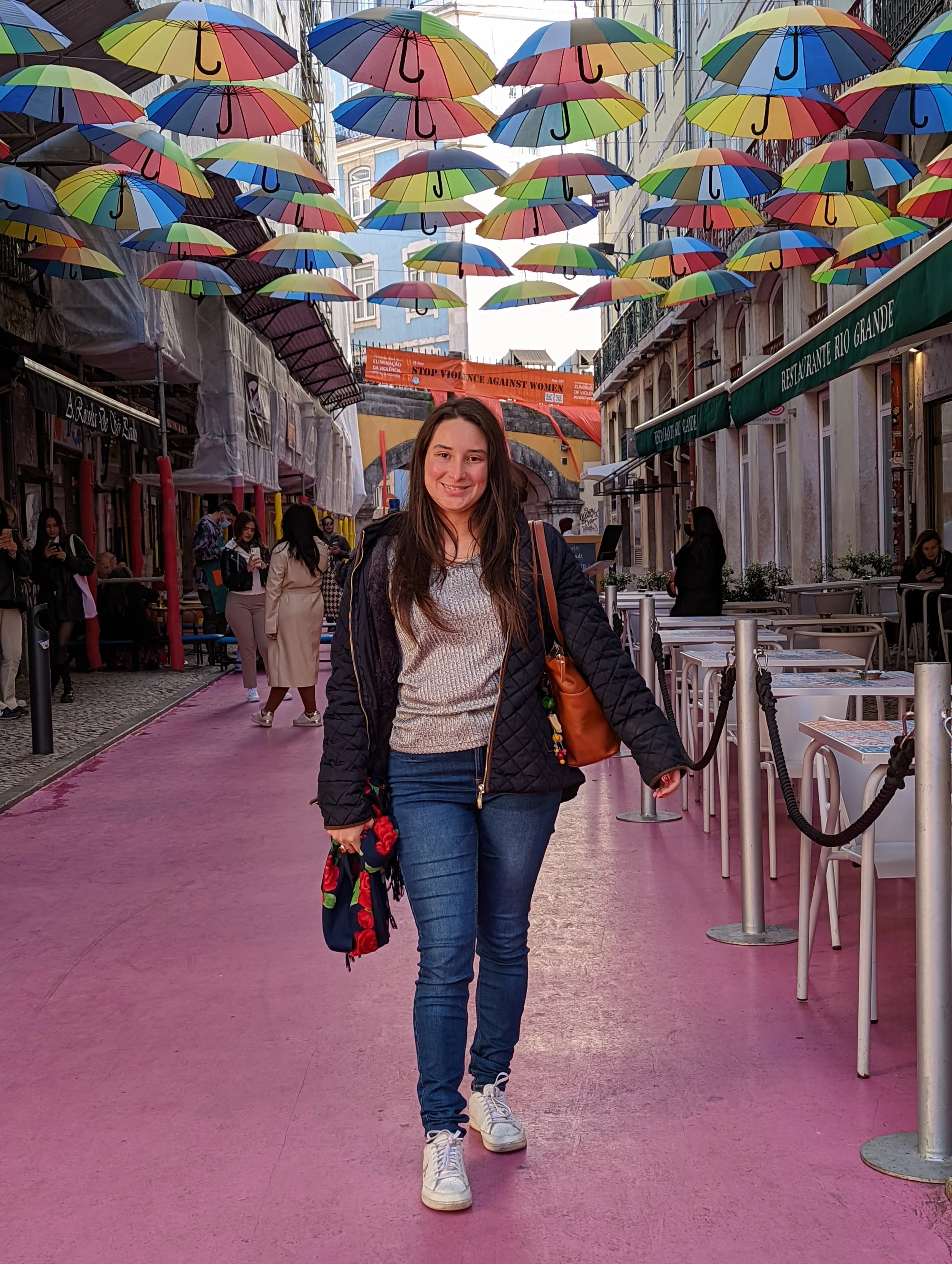 A young woman with long brown hair smiling while walking on a pink pedestrian street decorated with colorful umbrellas hanging above. She is wearing a beige sweater, blue jeans, sneakers, and carrying a brown bag.