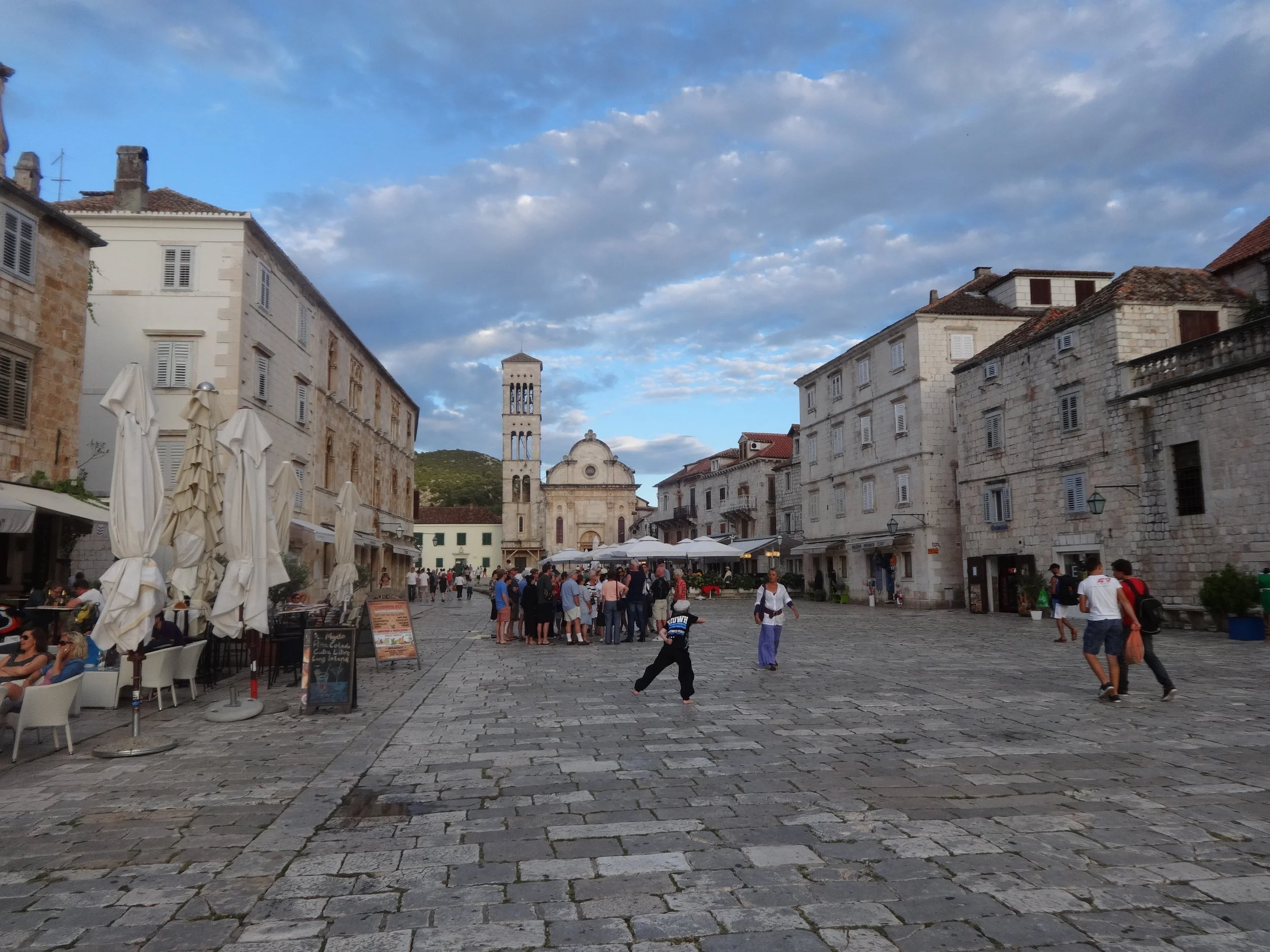 A lively European town square with people walking, sitting at outdoor cafes, historic stone buildings, and a church with a bell tower. The sky is partly cloudy.