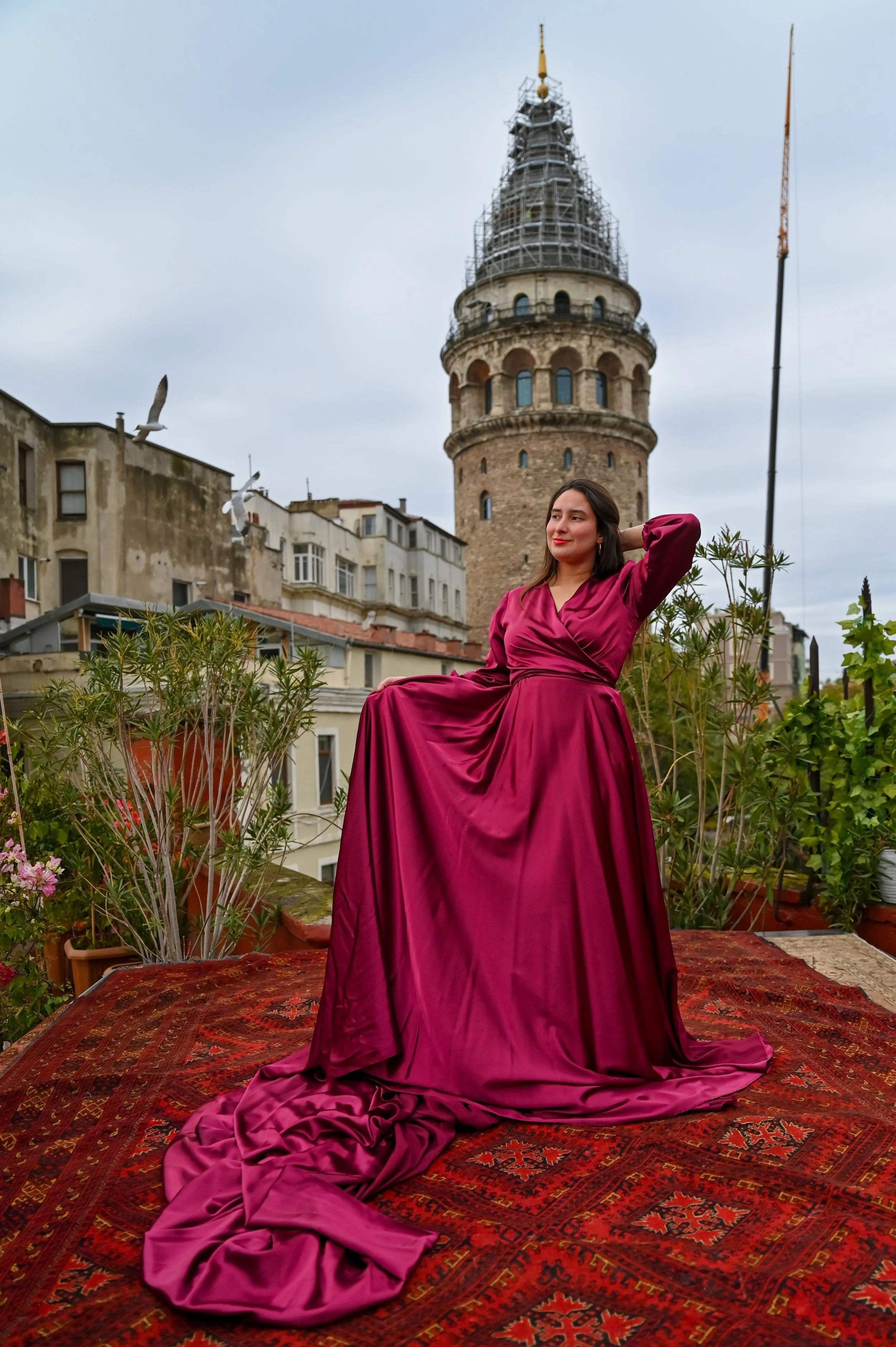 A woman in a deep red, satin, floor-length gown sitting outdoors on a patterned rug with plants in the foreground. Behind her are old buildings and a historic tower under a cloudy sky.