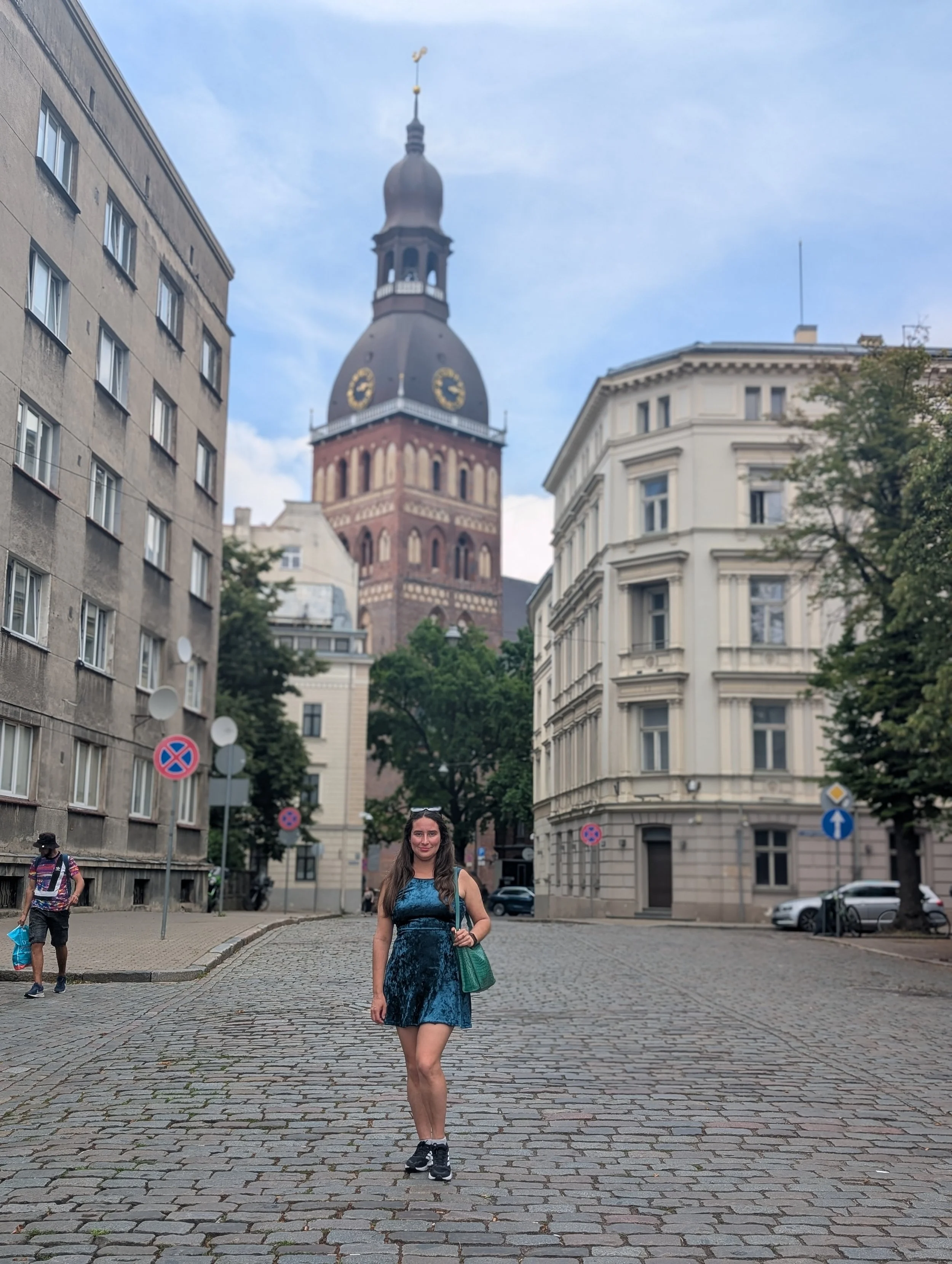 A woman wearing a blue dress and black sneakers walking on a cobblestone street in an urban area with historic buildings and a clock tower in the background.