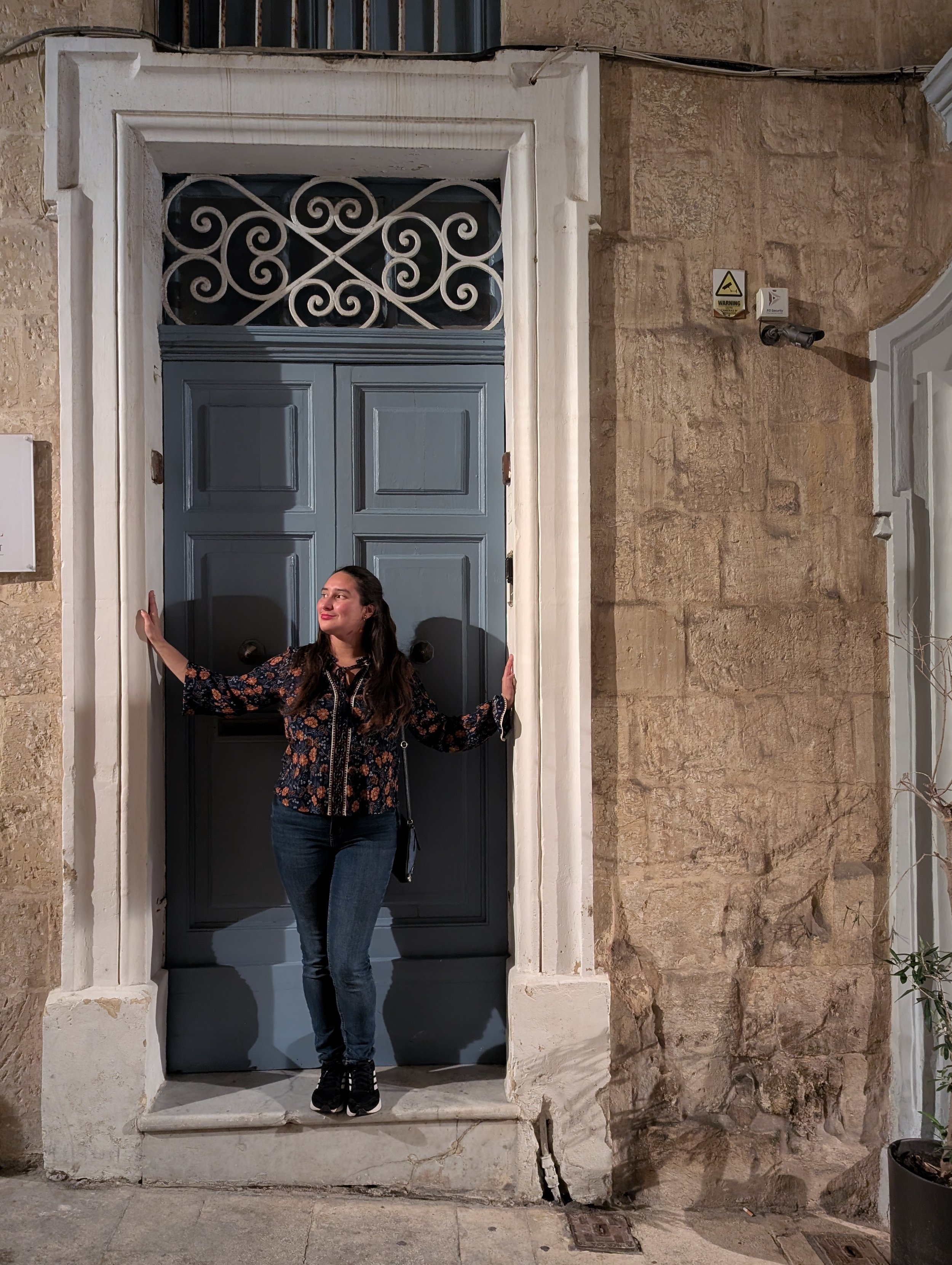 A woman standing at a large blue door with her arms outstretched, in front of a stone wall and security camera, at nighttime.