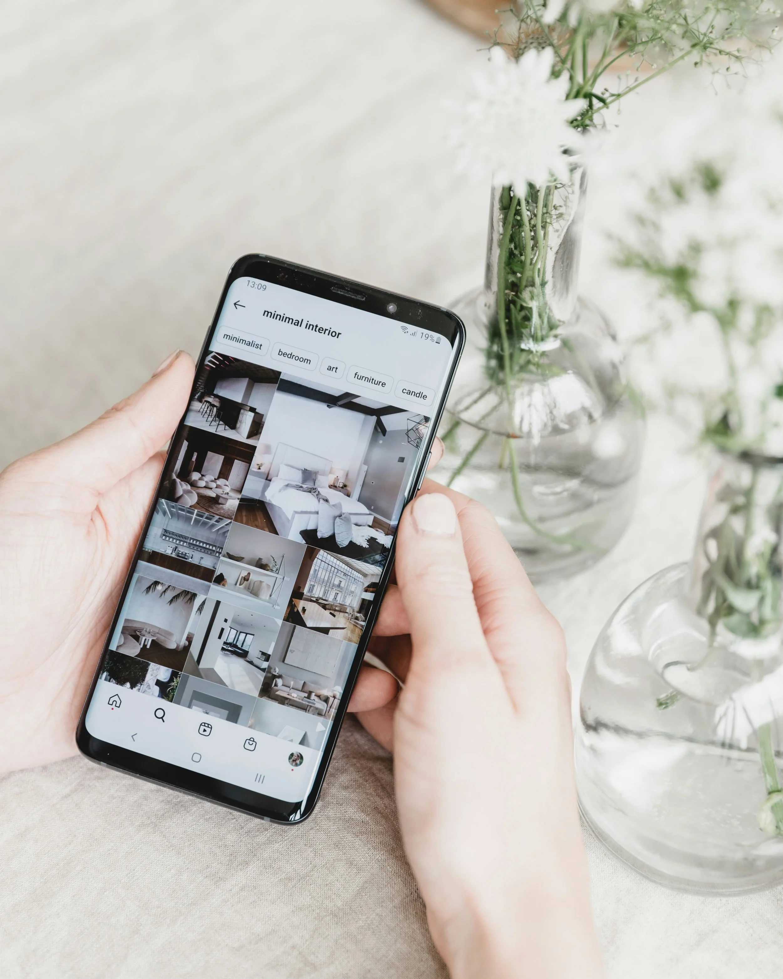 Hands holding a smartphone displaying minimalist interior design photos beside a glass vase of white flowers.