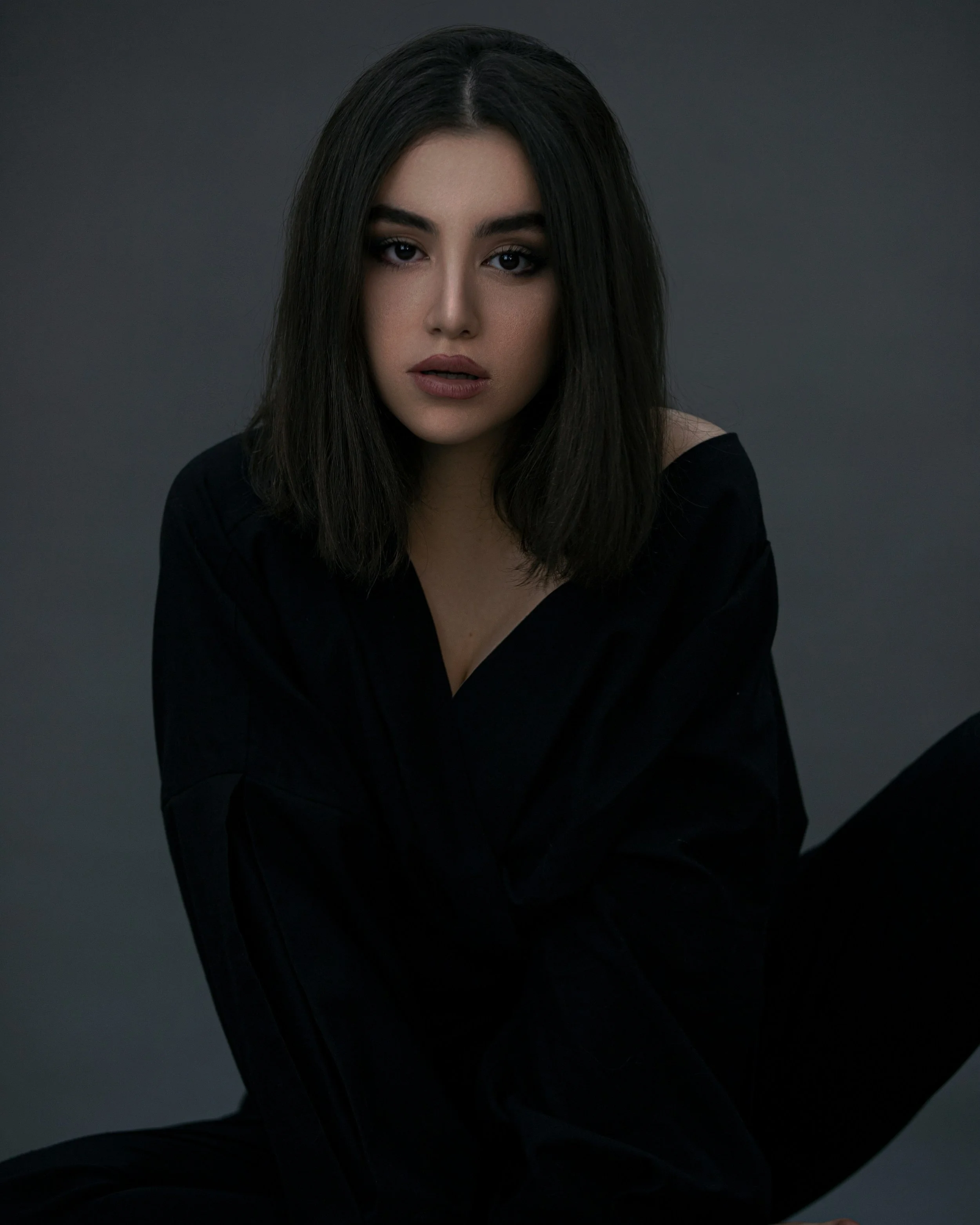 Close-up portrait of a woman with dark hair and dramatic eye makeup wearing a black top against a gray background.