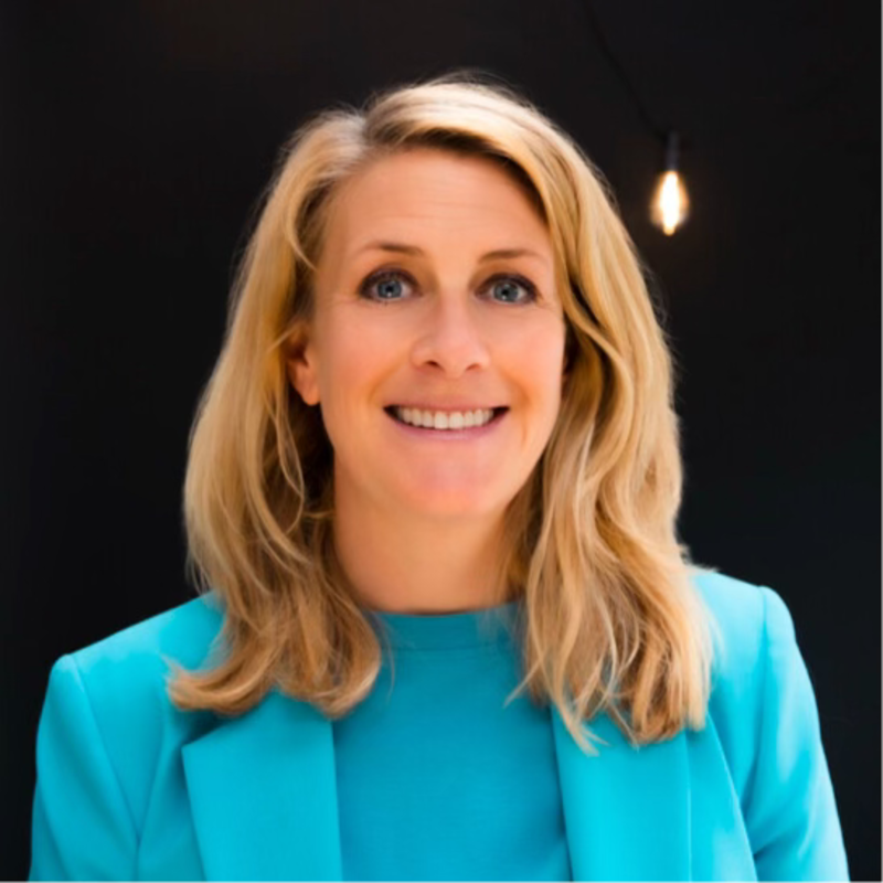 Smiling blonde woman in a turquoise blazer against a dark background
