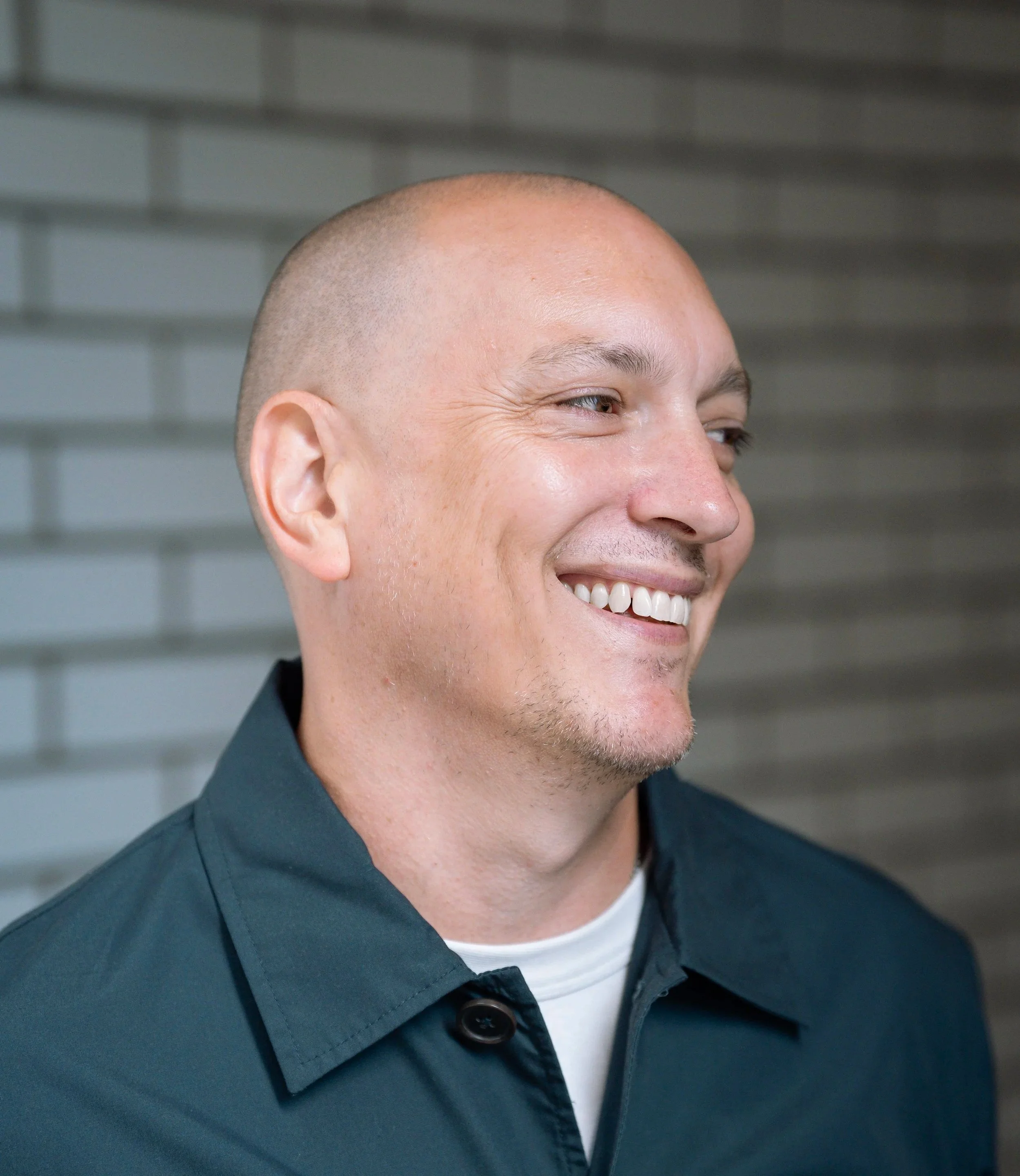 Close-up portrait of a smiling bald man with light stubble wearing a dark jacket