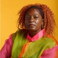 Portrait of a woman with curly reddish-brown hair wearing a bright pink shirt and multicolored green and orange scarf