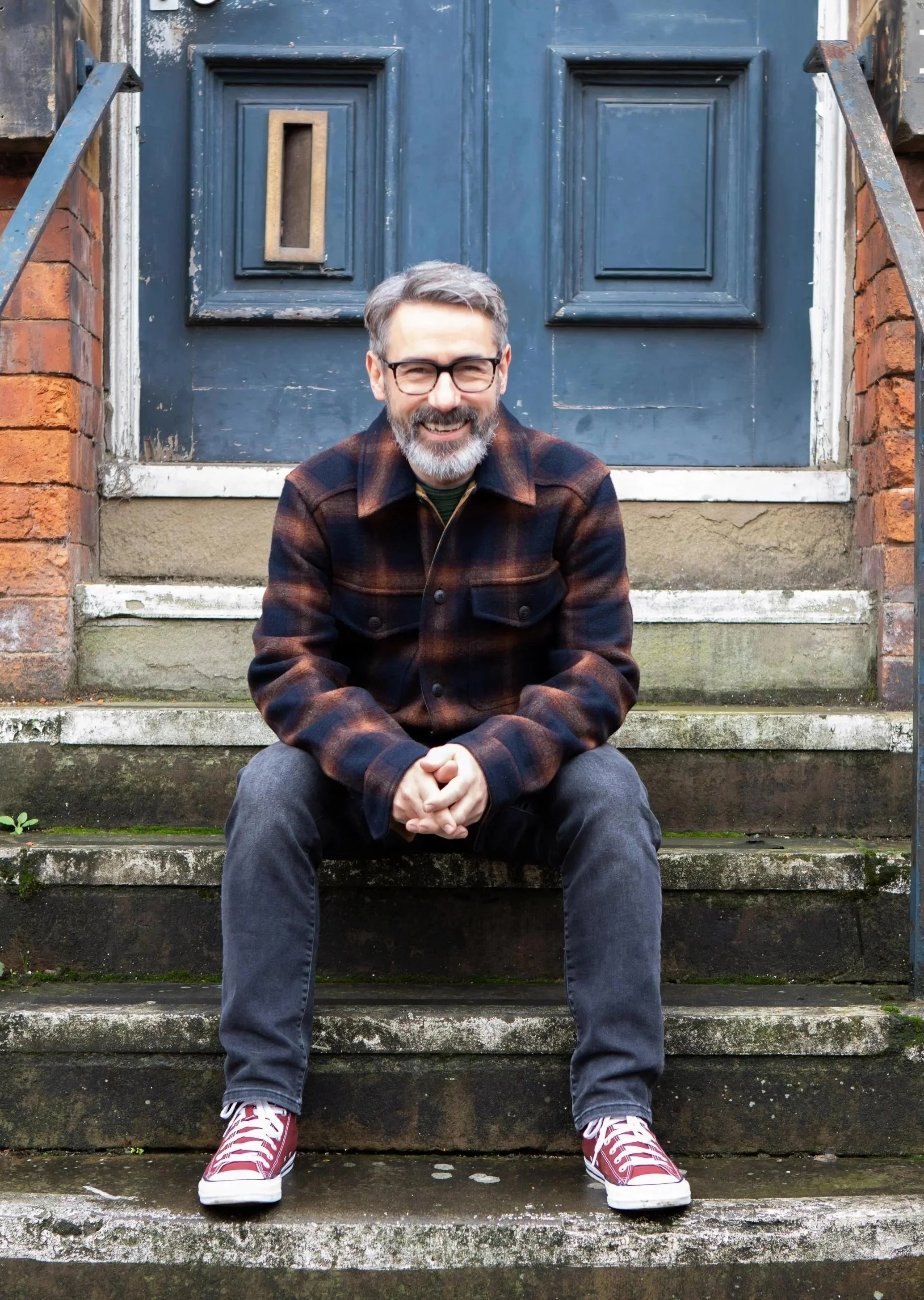 Smiling bearded man wearing glasses and a plaid jacket sits on stone steps in front of a blue door.