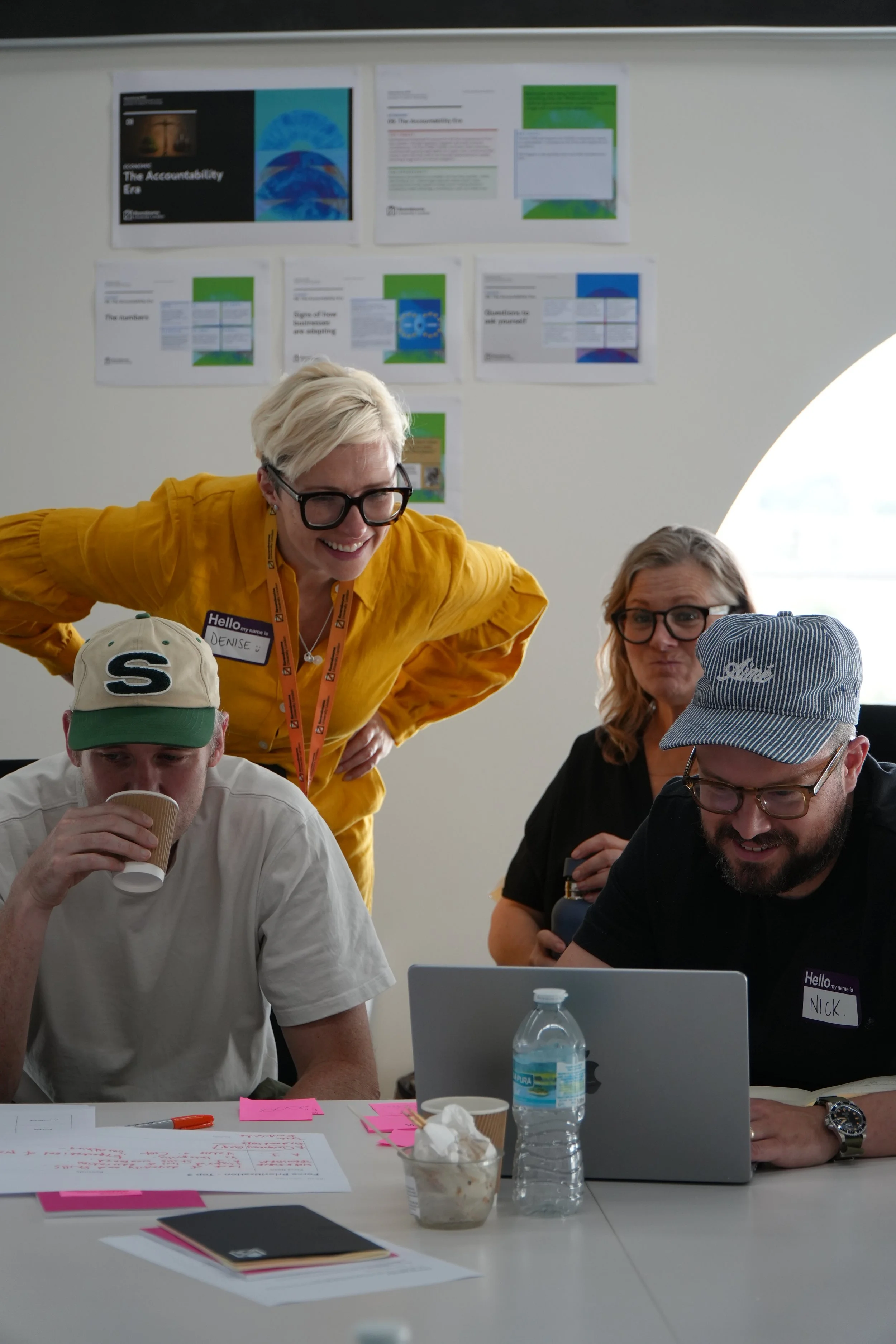 People collaborating around a table with a laptop, coffee, water bottle, and workshop notes beneath presentation posters.