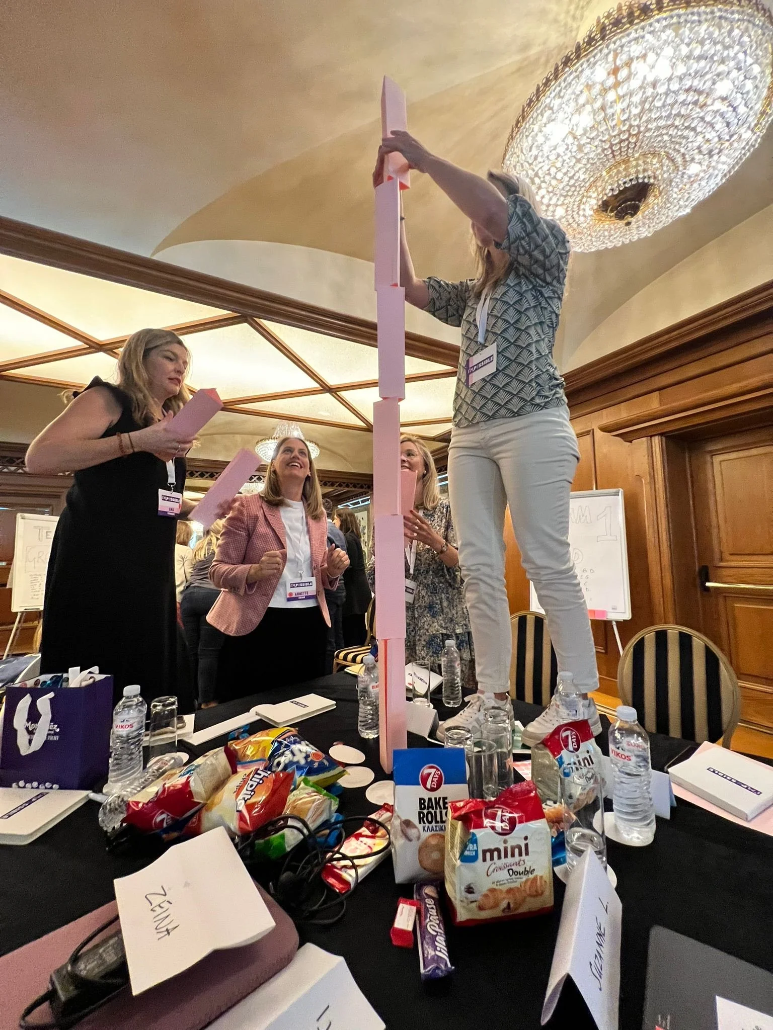 Women at a workshop building a tall pink tower from paper strips around a snack-covered conference table.