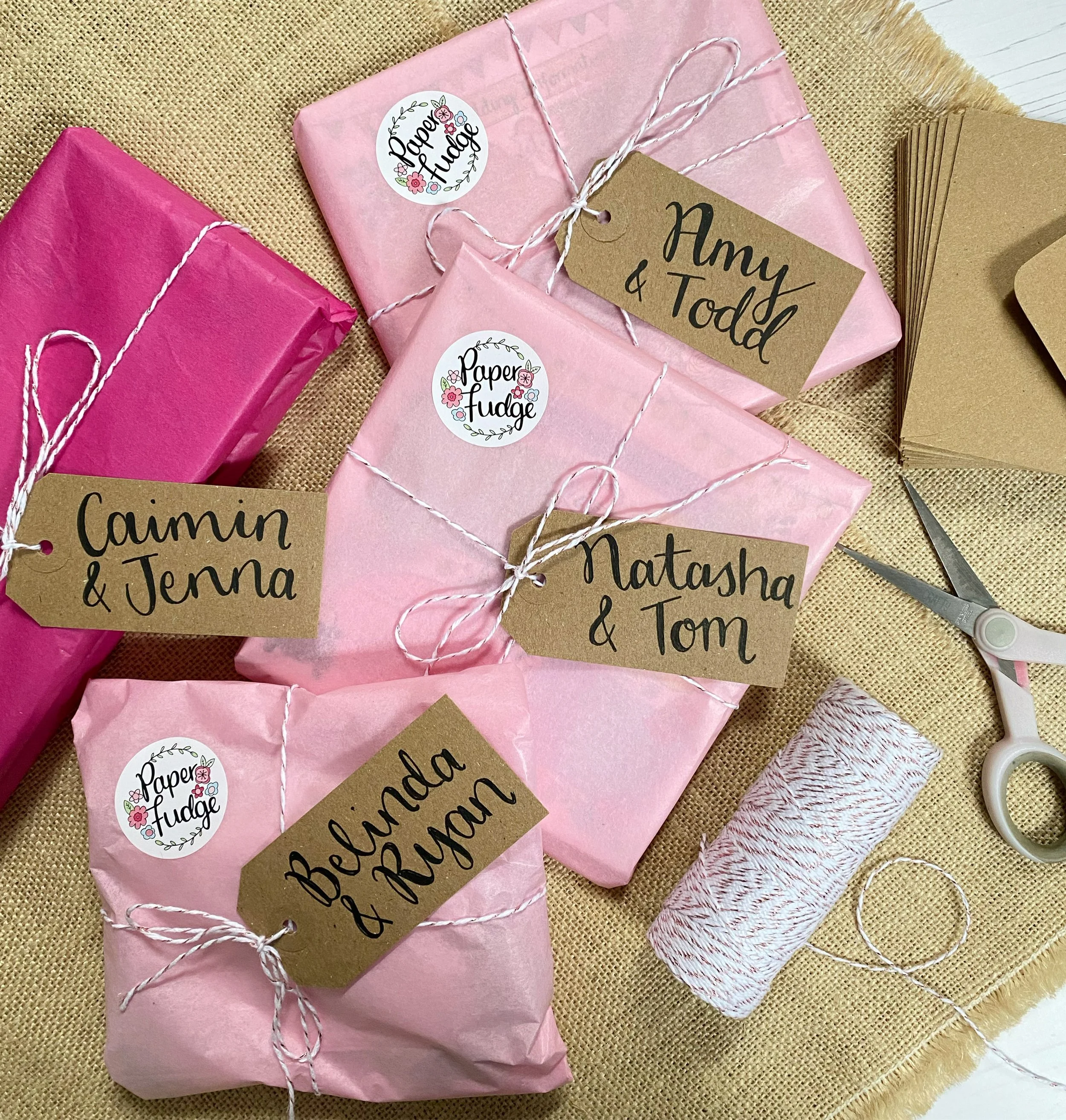 Multiple wrapped gifts on a beige surface, pink paper, brown 'Amy & Todd' tags, and stickers reading 'Paper Fudge' with floral designs.