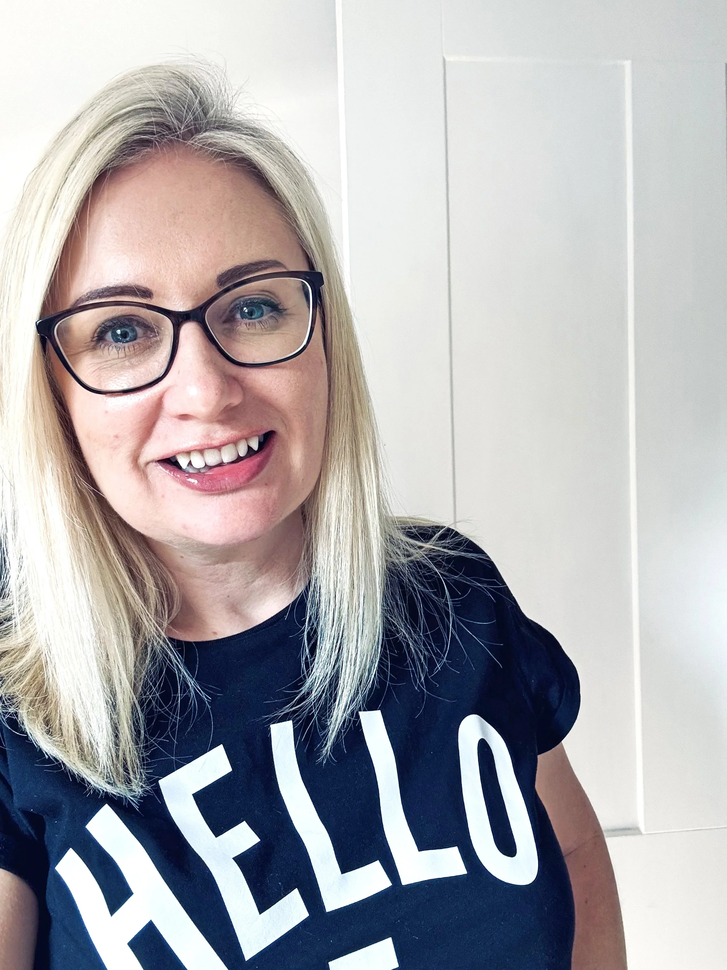 Woman with blonde hair wearing black glasses and a black t-shirt with white text.