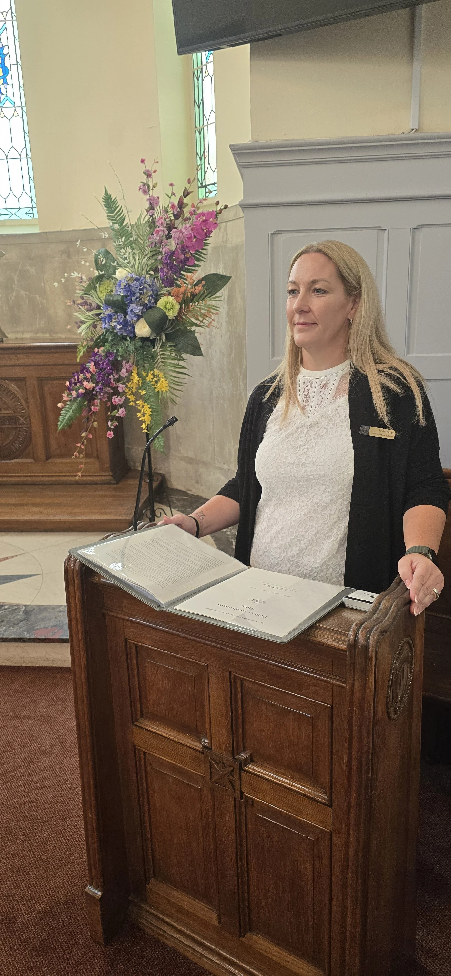 A woman standing behind a wooden stand with open folders, inside a church with stained glass windows and a colorful flower arrangement to her side.