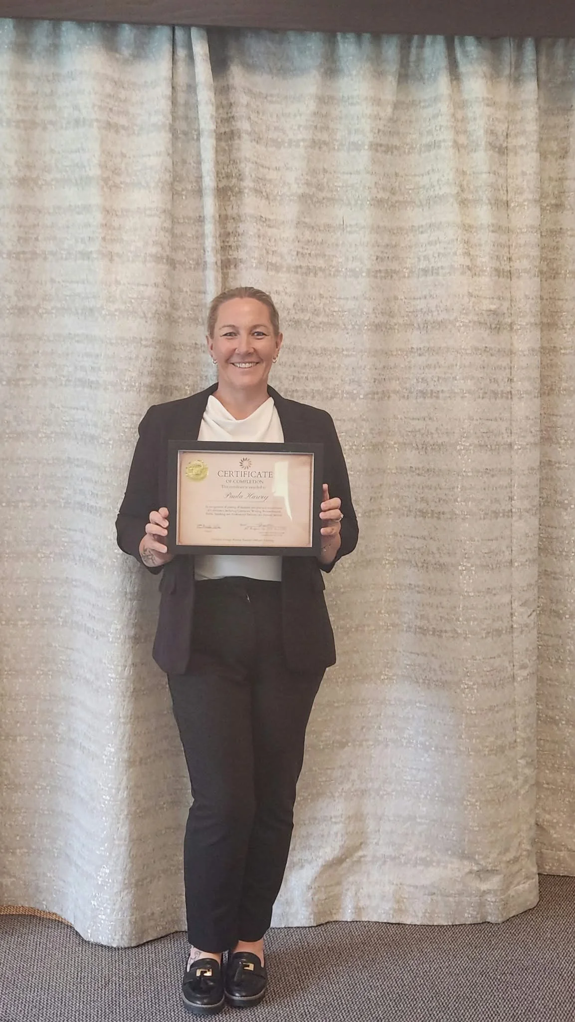 A woman in a black blazer and pants holding a framed certificate, standing in front of beige curtains, smiling at the camera.
