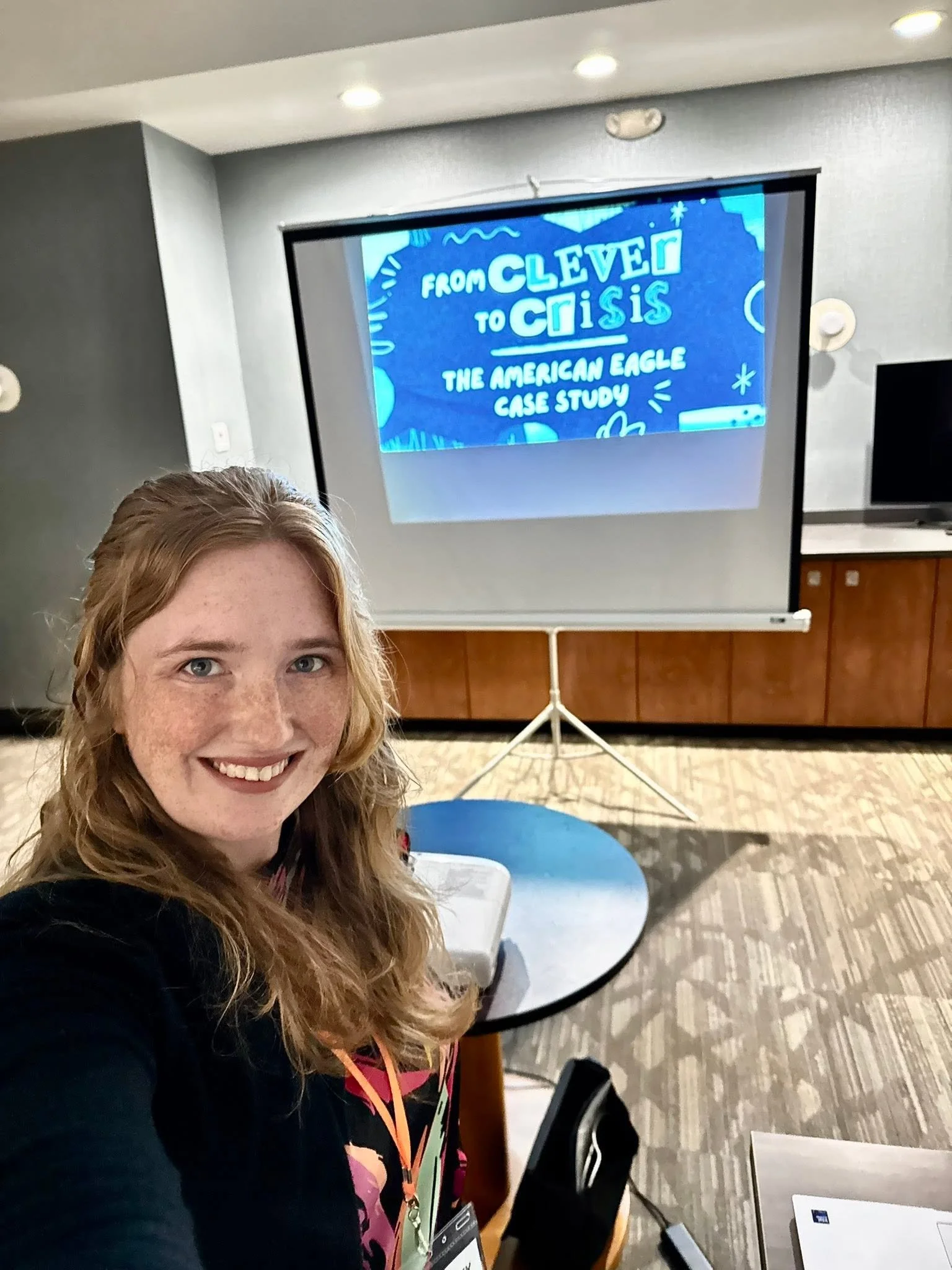A woman with red hair and freckles smiling for a selfie in front of a projection screen displaying a presentation titled 'From Clever to Crisis: The American Eagle Case Study', at a conference or seminar room.