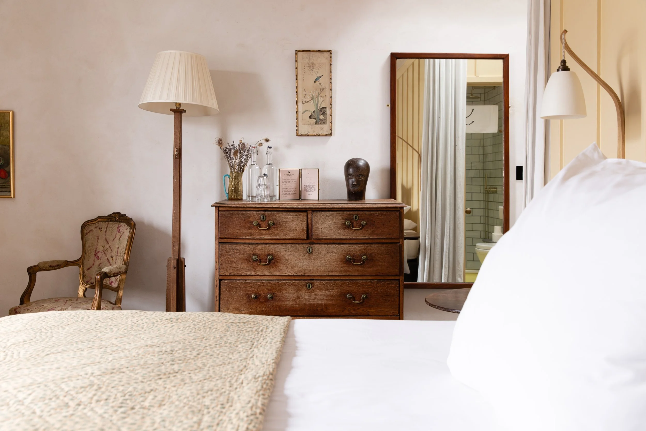 A bedroom with a bed in the foreground, a vintage chair, a wooden dresser with decorative items, and a doorway to a bathroom with shower tiles.