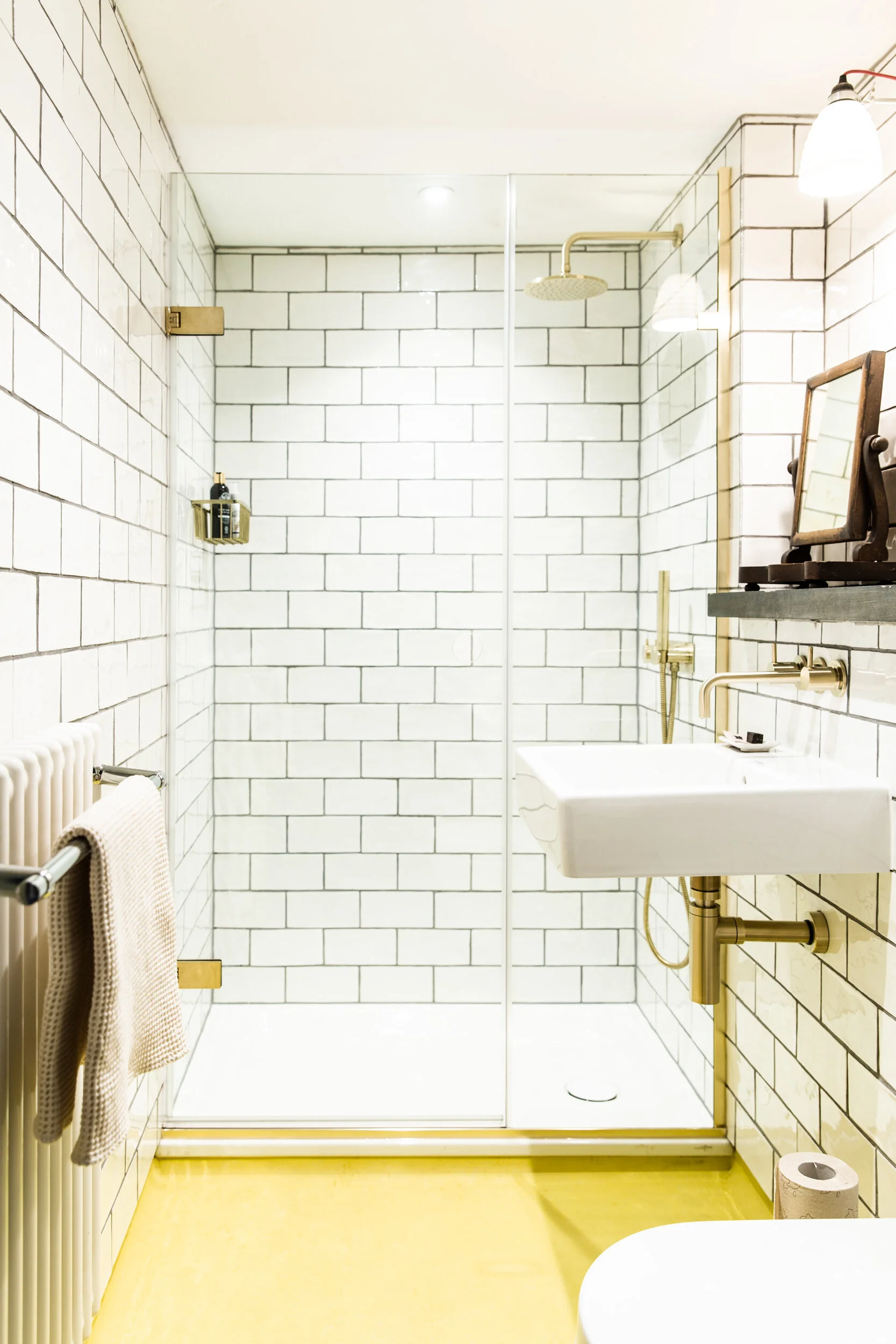 Bathroom with white subway tiles on the walls, yellow floor, and a glass shower enclosure. There is a wall-mounted sink with a mirror above it, brass fixtures, and a toilet with a roll of toilet paper nearby. A towel hangs on a radiator, and a small 