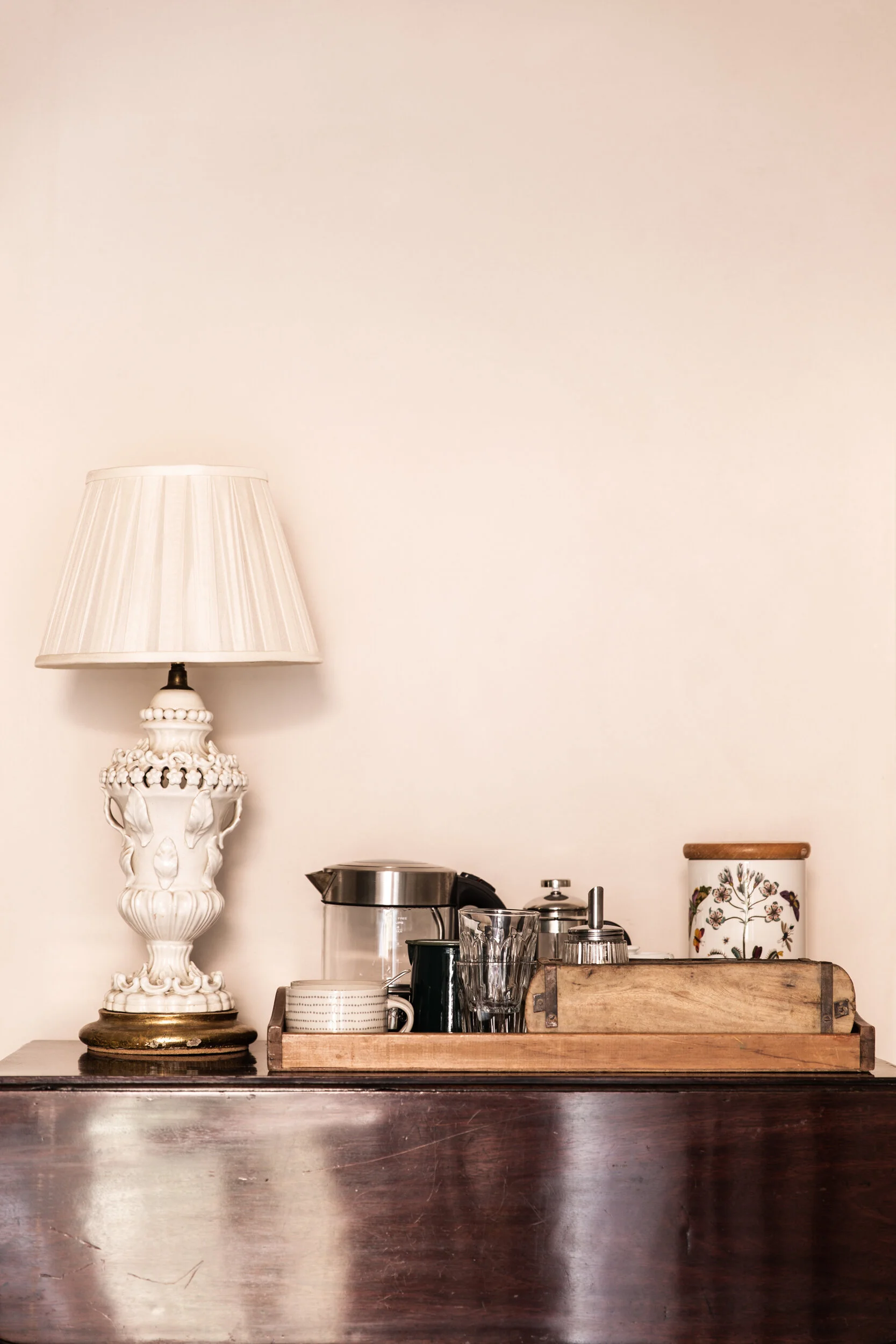 A cream-colored table lamp with a decorative ceramic base and a pleated shade. The lamp is on a countertop next to kitchen utensils and a wooden tray.