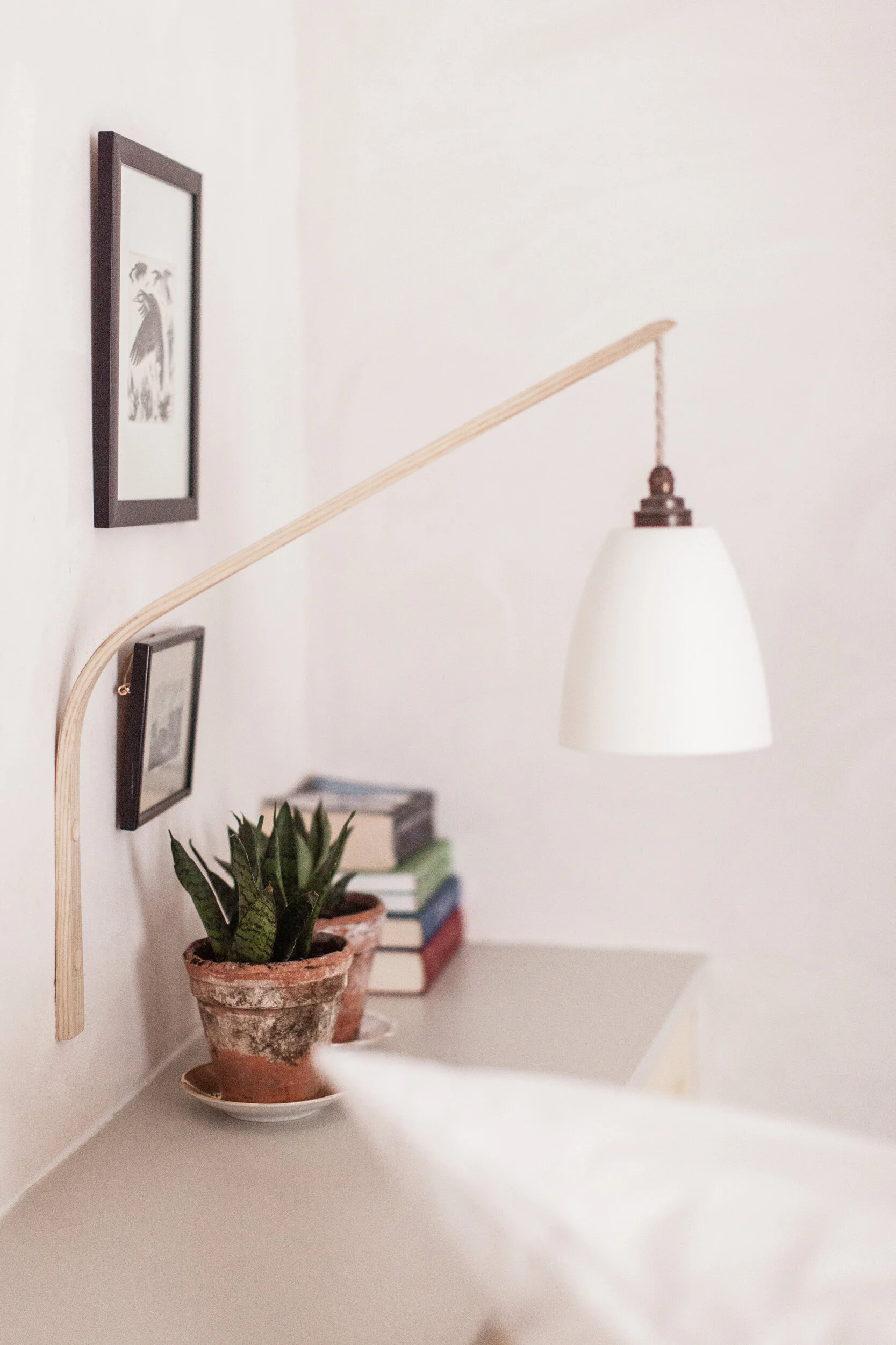 Close-up of a cozy corner with a white desk, potted plant, stacked books, framed pictures on the wall, and a white hanging light fixture.