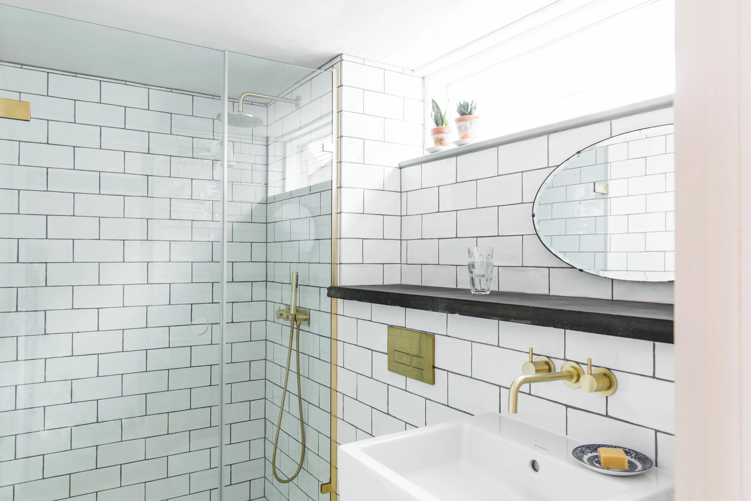 Modern bathroom with white subway tile walls, a glass shower enclosure, a black countertop with a glass of water, a soap dish with soap, and potted plants on a windowsill.