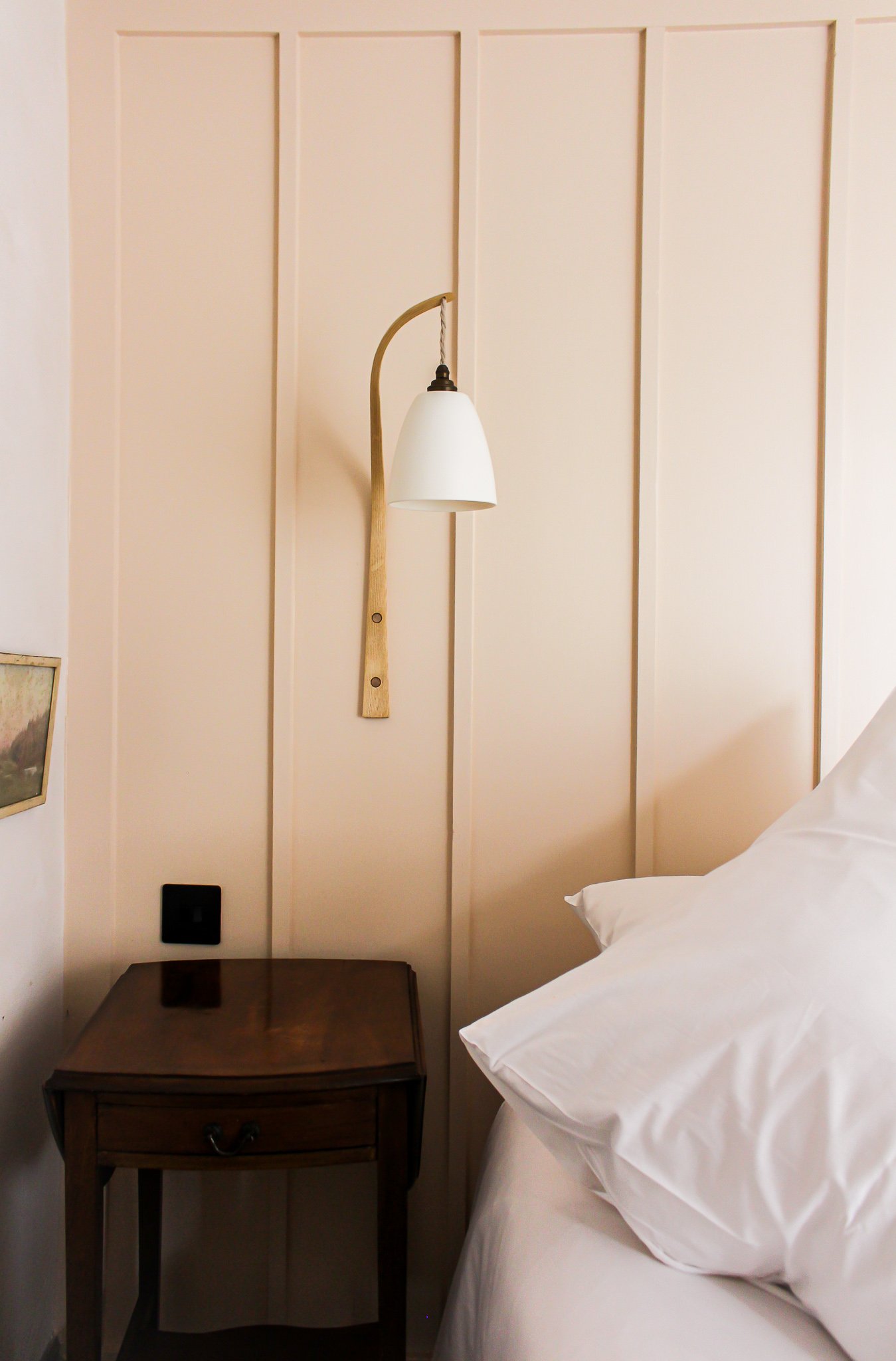 A bedside wall with a wooden headboard, a small wooden nightstand, a bed with white sheets and pillows, a wall-mounted reading lamp, and a framed picture on the wall.