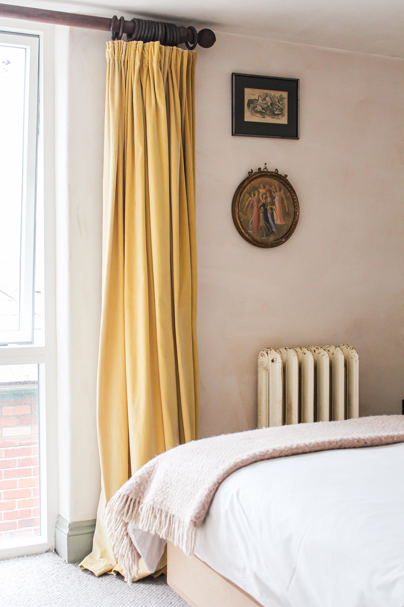 A bedroom corner with a window framed by beige curtains, a vintage framed painting of ducks, a decorative round religious painting of angels, a white radiator, and part of a bed with a beige blanket.