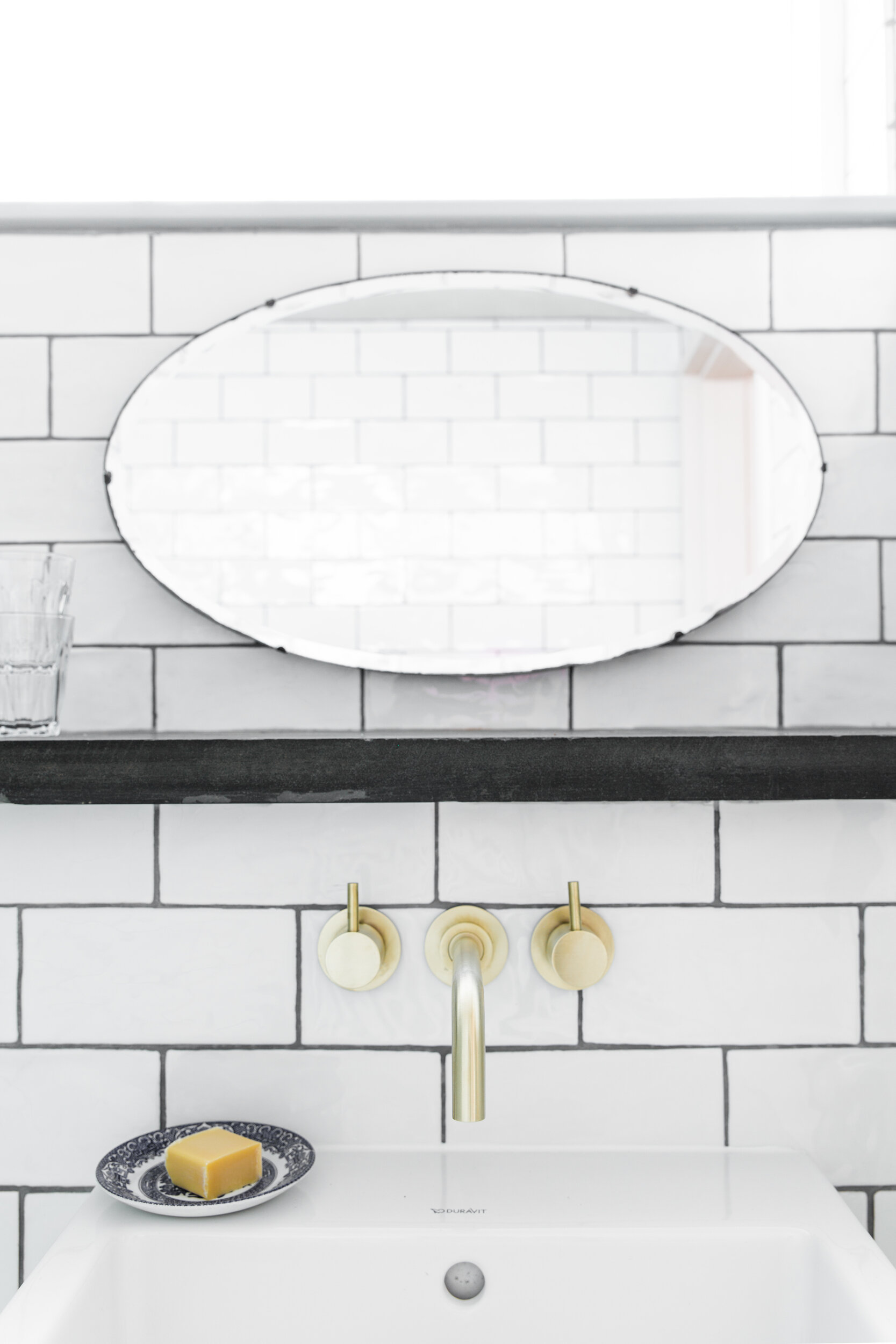 Modern bathroom with white tiled walls, a white sink, a large oval mirror, a gold faucet, three cream-colored soap dispensers, a decorative plate with soap, and a window in the background.