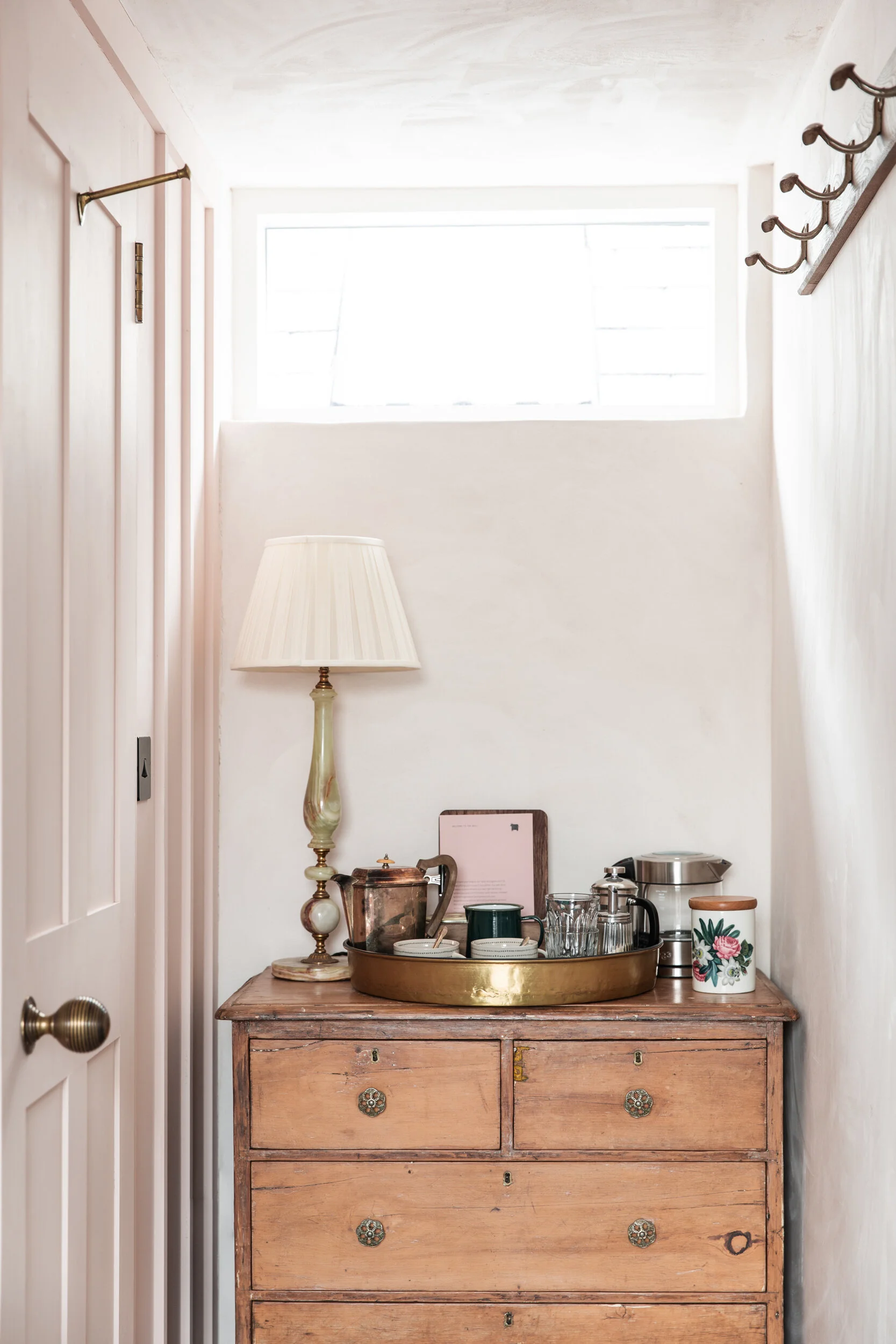 A wooden dresser with vintage drawer pulls holding various kitchen items, including a metal pitcher, a tray with cups, glasses, and a teapot. To the left, a tall vintage-style table lamp with a fabric shade stands. The background features a white wal