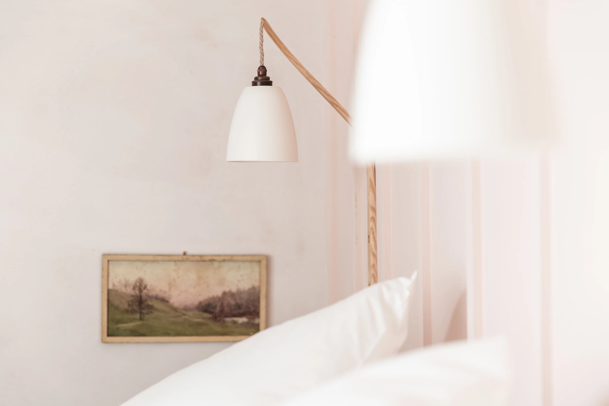 A minimalist bedroom with a white wall, a framed landscape photograph, and a wooden and white wall-mounted lamp.