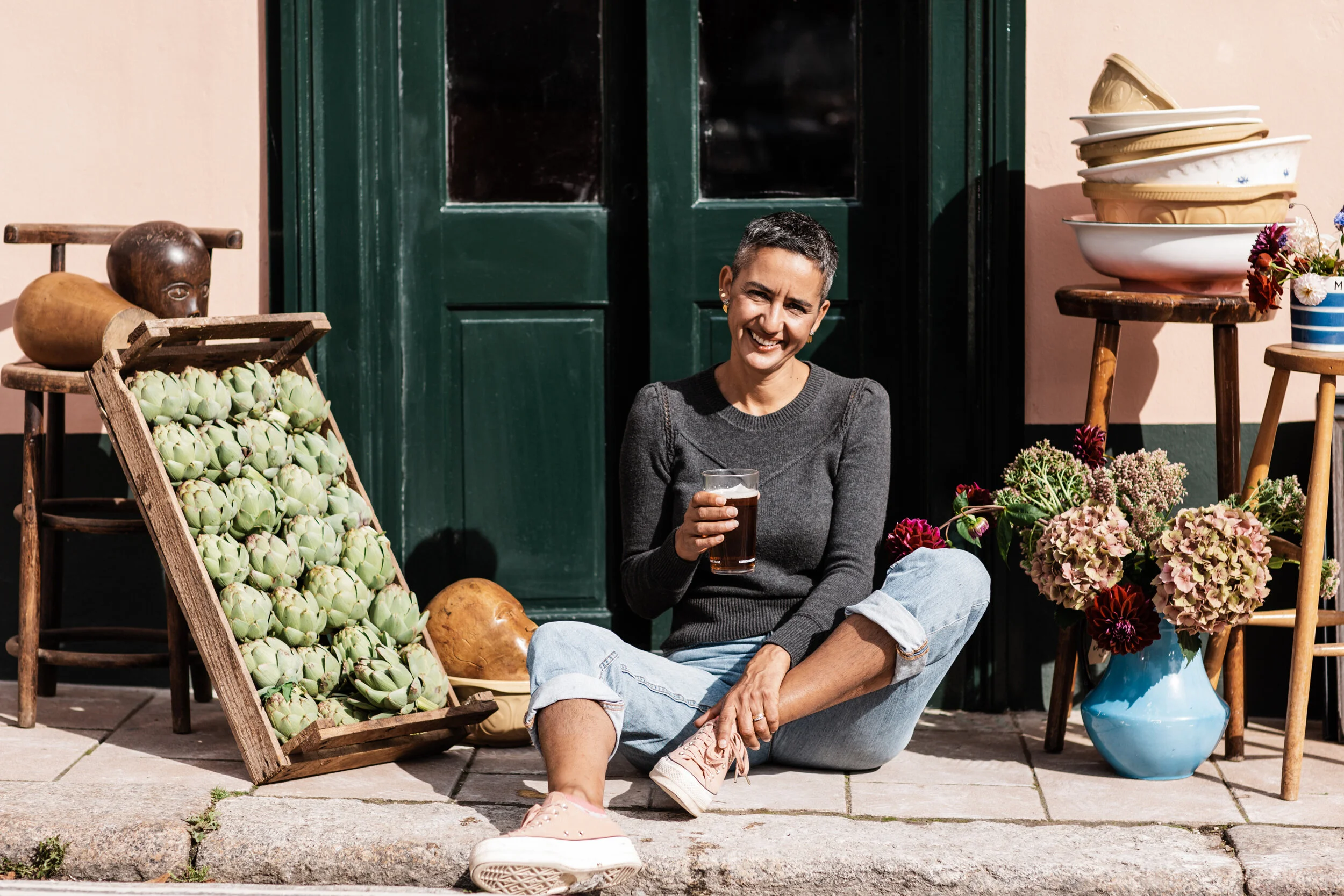 A woman with short gray hair and earrings, sitting on the sidewalk in front of a green door surrounded by flower arrangements and pottery, holding a glass of dark beverage, smiling.