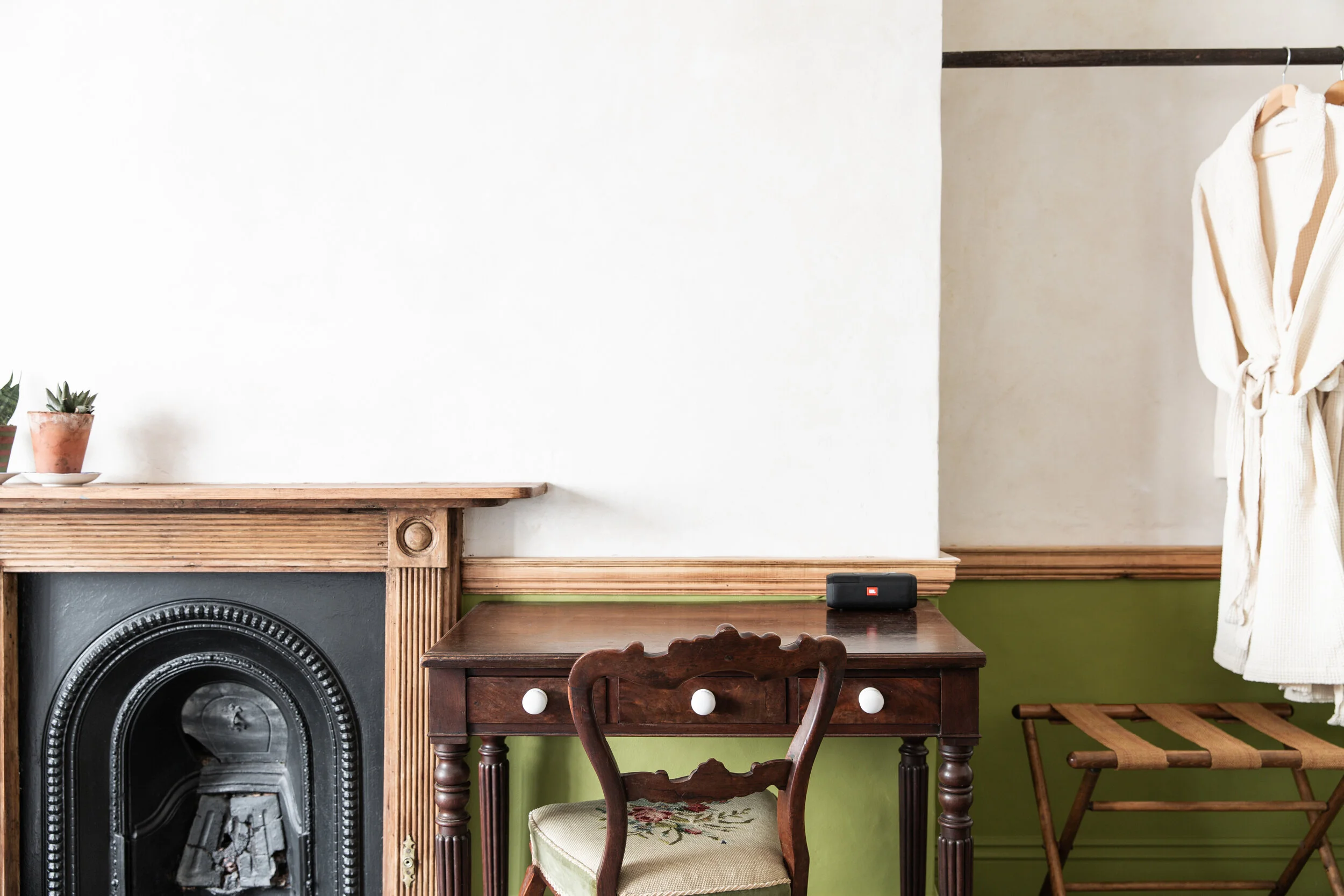 A cozy room corner with a fireplace with a wooden mantel, a small antique wooden desk, a vintage wooden chair with a cushioned seat, and a small luggage rack, against a green and beige wall with a curtain on the right.